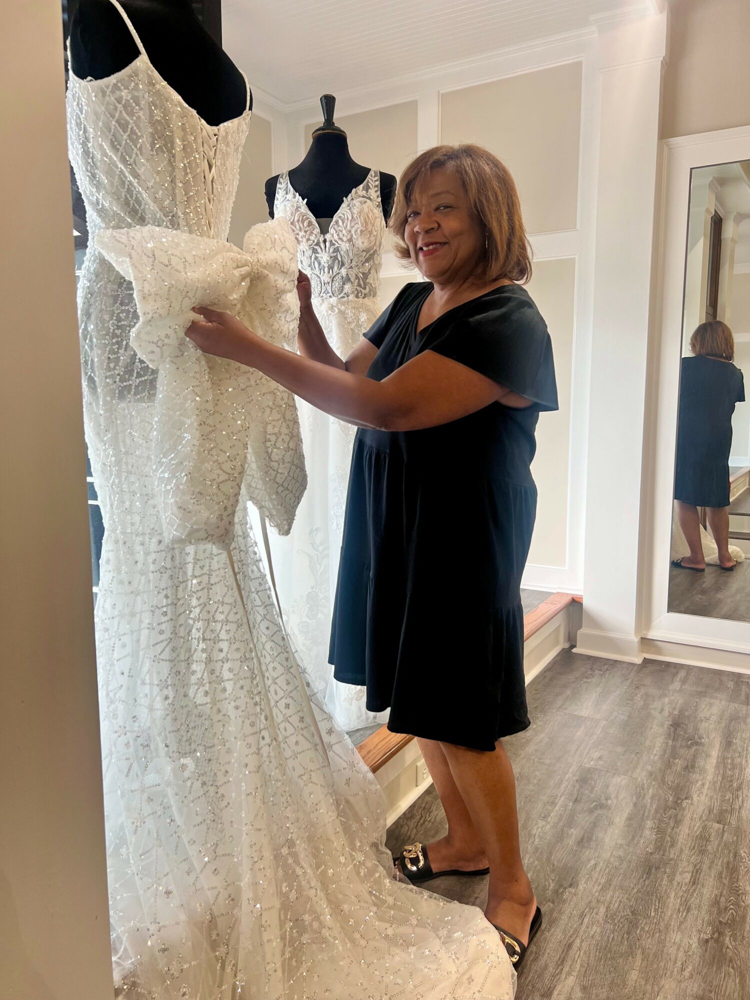 Woman with shoulder-length hair in a black dress stands next to a mannequin wearing a white lace wedding gown with a large bow at the waist, inside a fitting room.