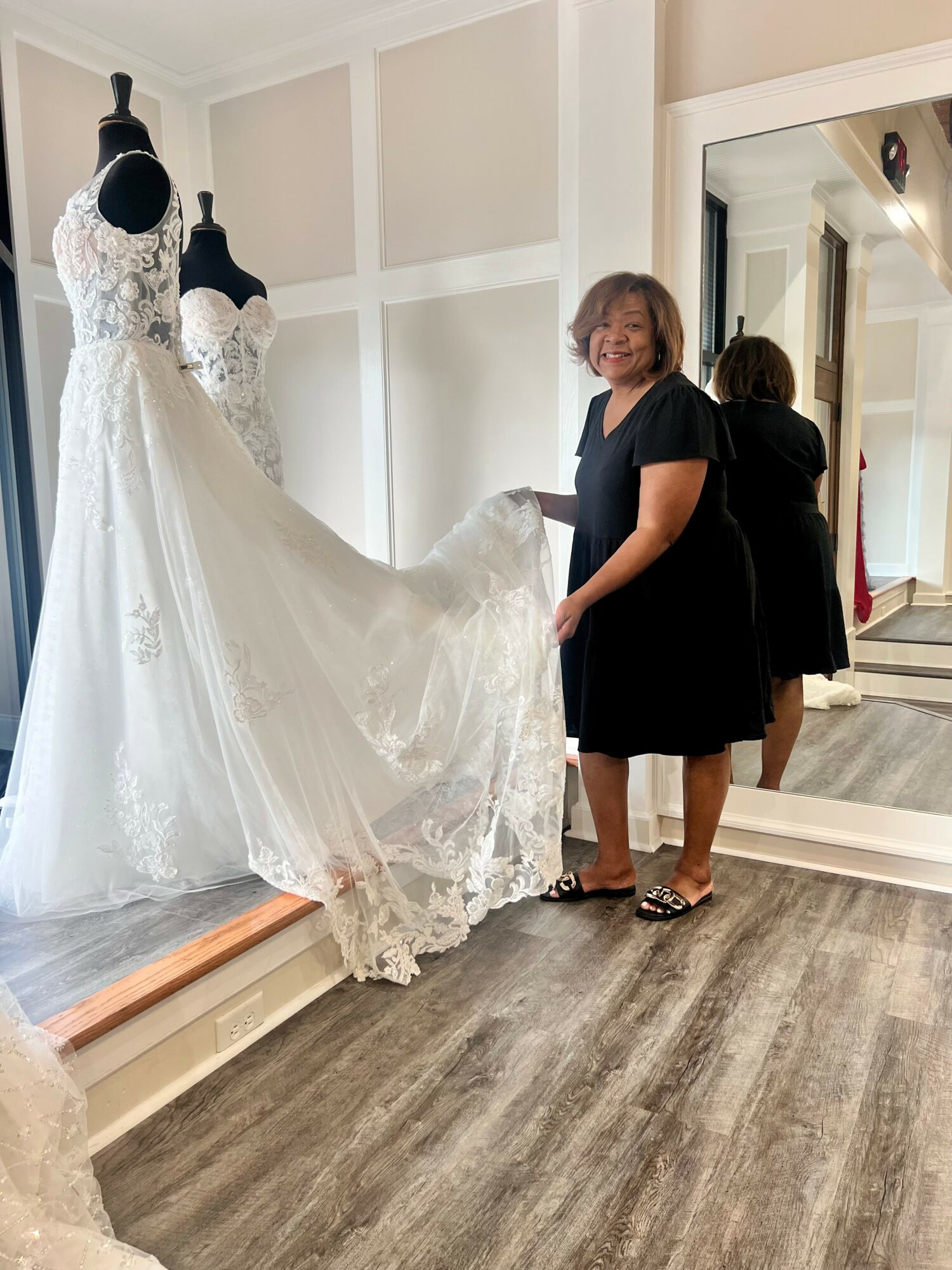 Woman in black dress holding a wedding dress on a mannequin, mirror reflects her and the dress, room with wooden floor.
