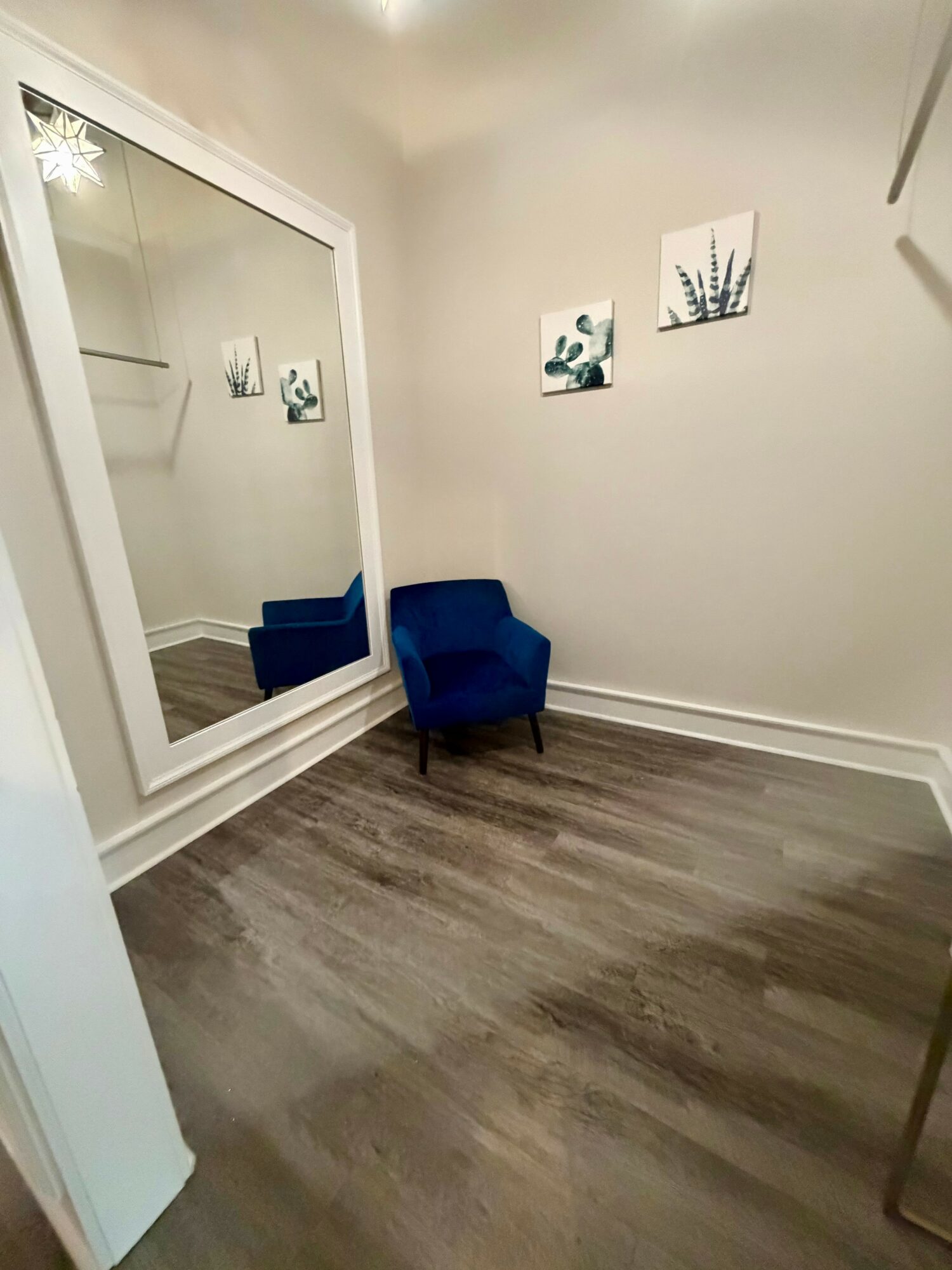 Corner of a room with a large mirror, a blue armchair, and three wall art pieces, with wood flooring.