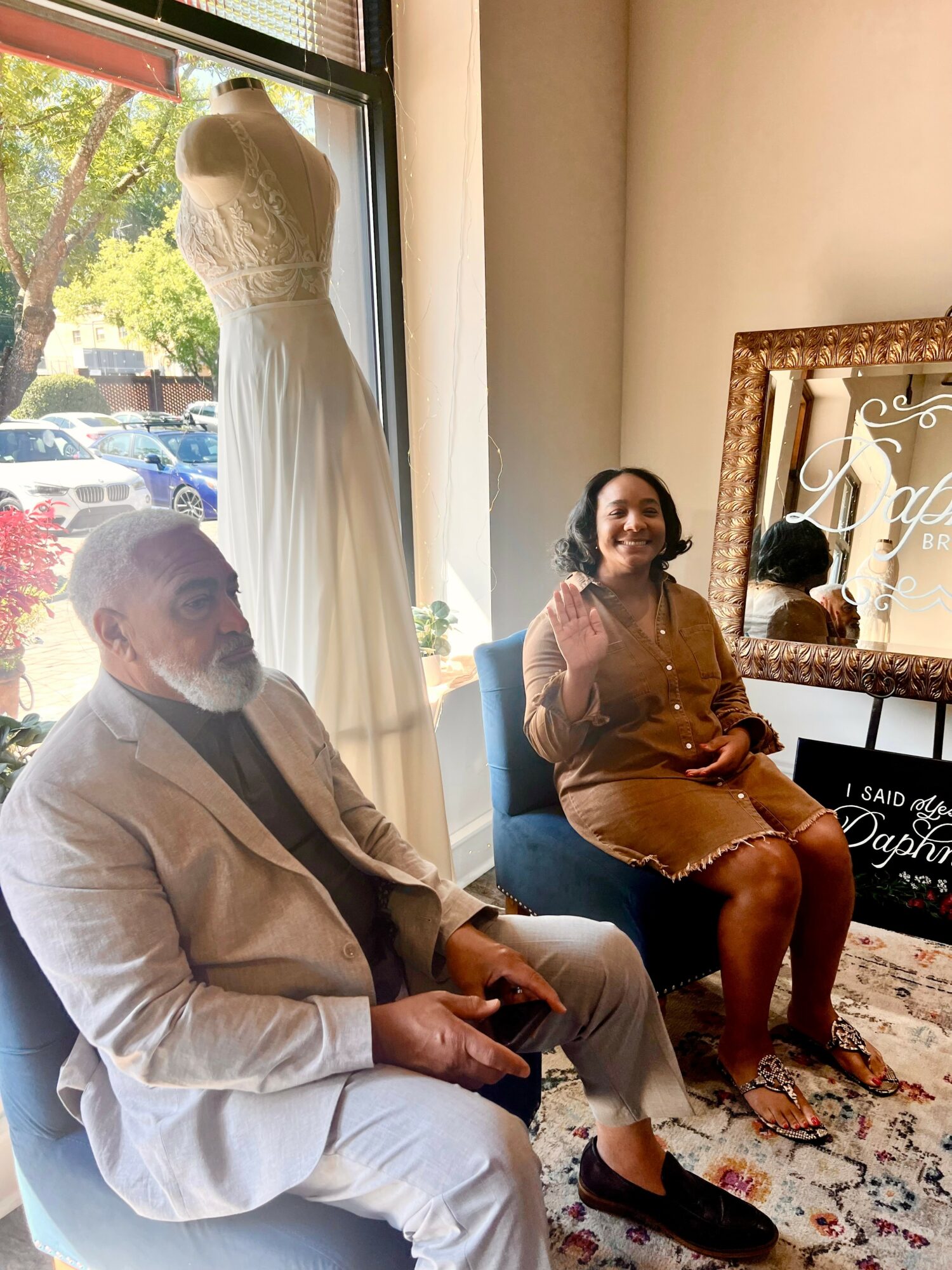 Two people sitting indoors near a window, with a dress on a mannequin behind them, and a mirror reflecting part of the room.