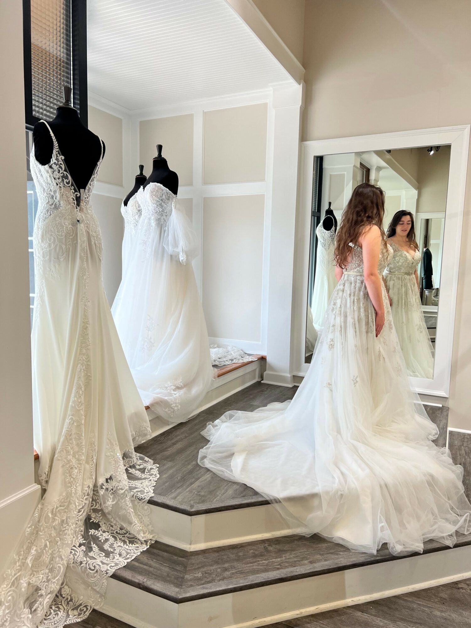 Person trying on a wedding dress in front of a mirror, with another wedding dress on a mannequin nearby.