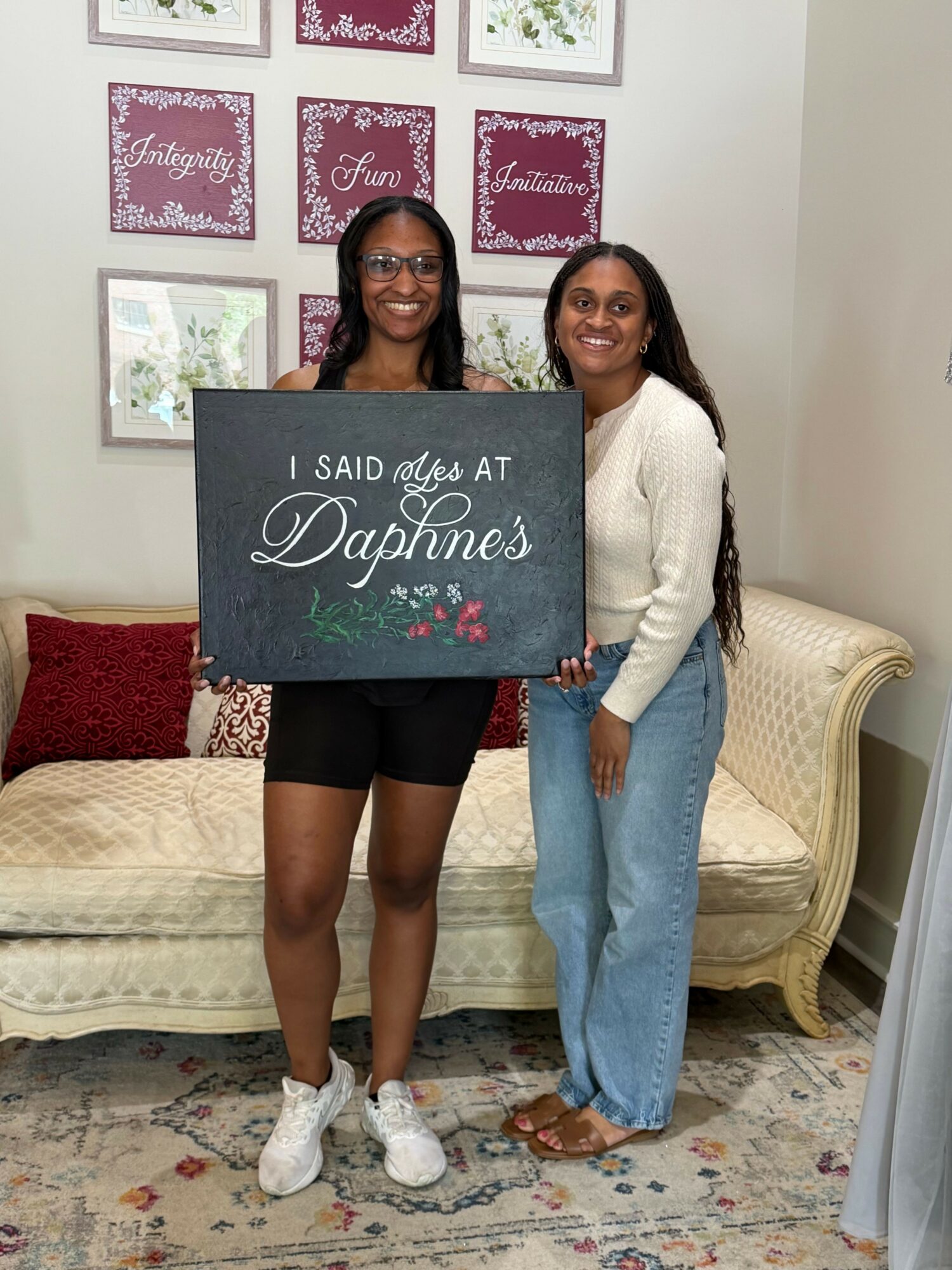 Two women standing in a living room, holding a sign that reads 'I said it at Daphnes,' with framed pictures on the wall behind them.