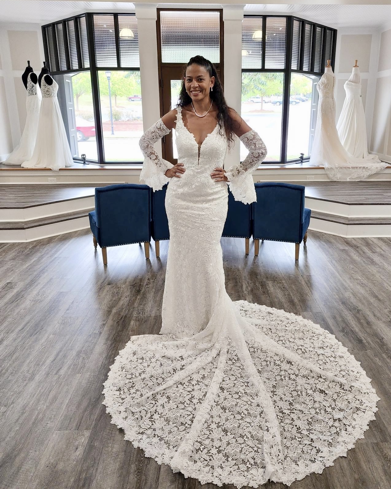 Woman in wedding dress standing in boutique with dresses on mannequins, large windows, and blue chairs behind her.