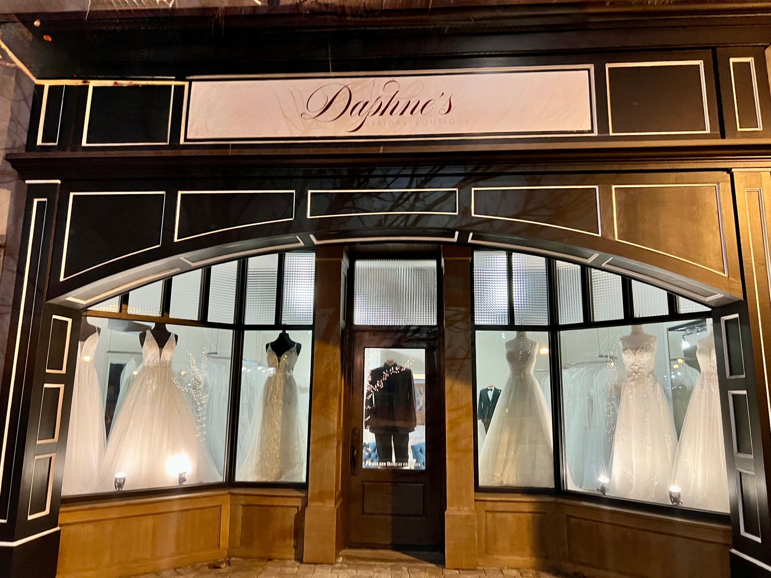 Storefront with five white dresses on mannequins in large display windows, sign above reads 'Daphne'.
