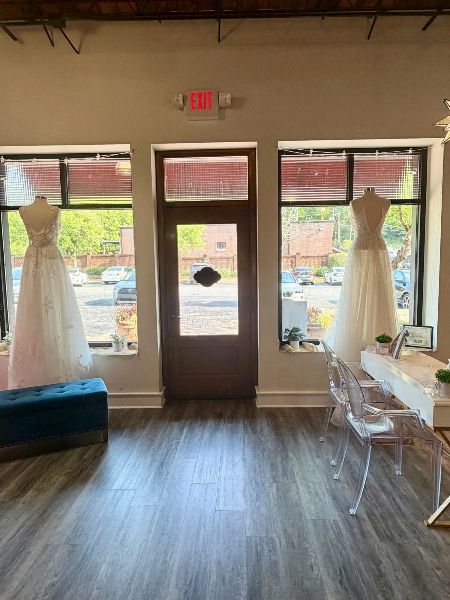 Interior view with a door between two large windows, two wedding dresses on mannequins, and a table with chairs.