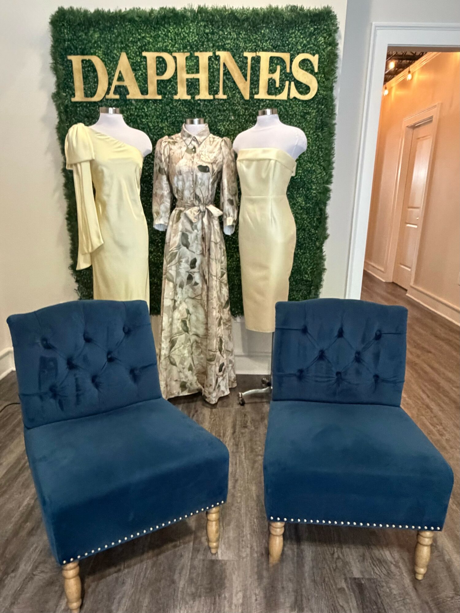 Three mannequins display dresses in front of a green wall with gold letters spelling 'DAPHNES'. Two blue chairs are in foreground.