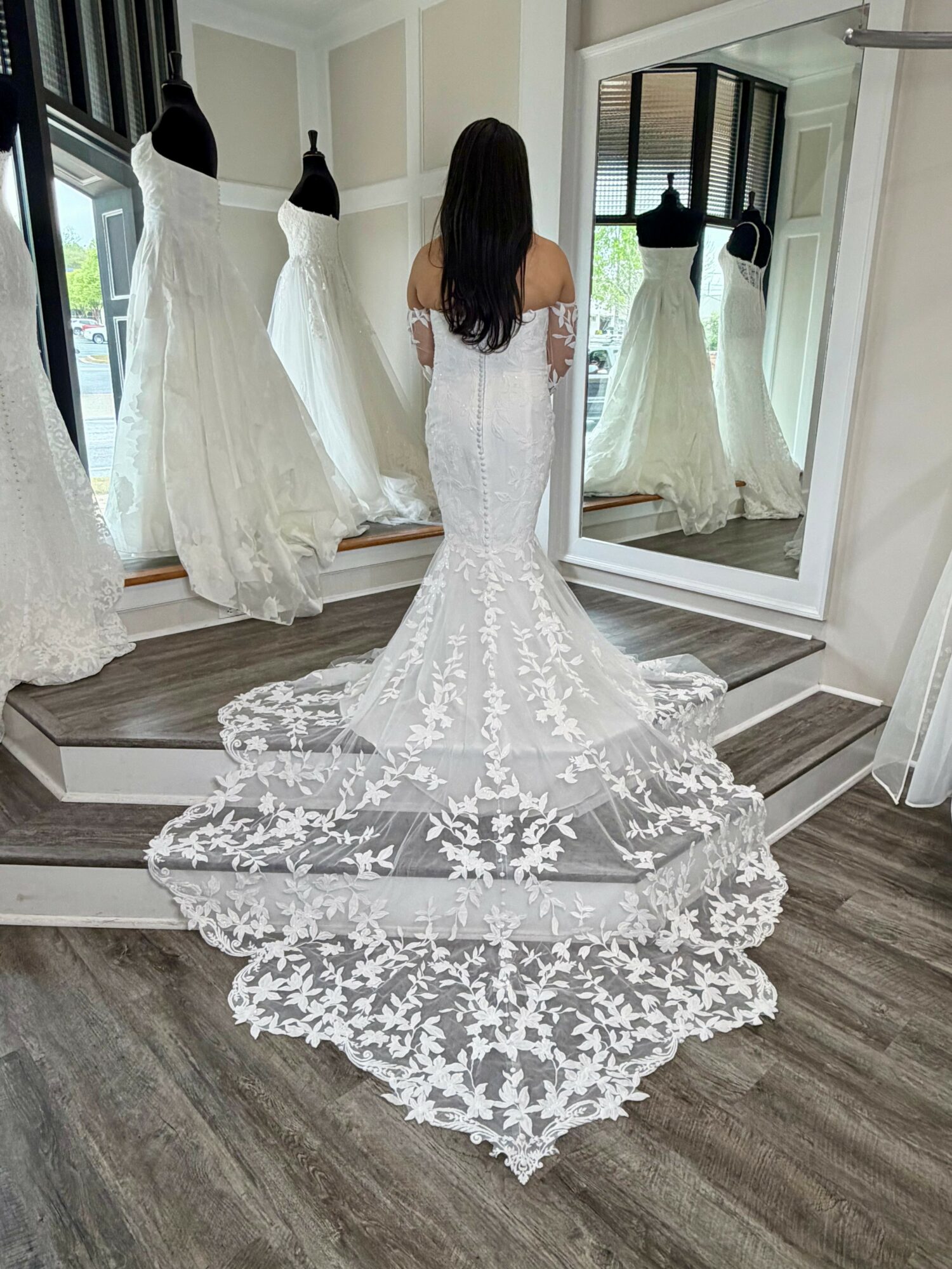 Woman in wedding dress looking at her reflection in a mirror, with three mannequins displaying dresses nearby.