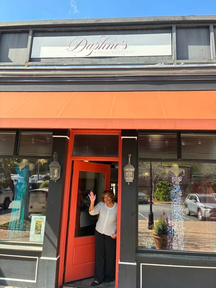 Person standing in doorway waving, with large windows and potted plants outside, storefront with orange awning above.