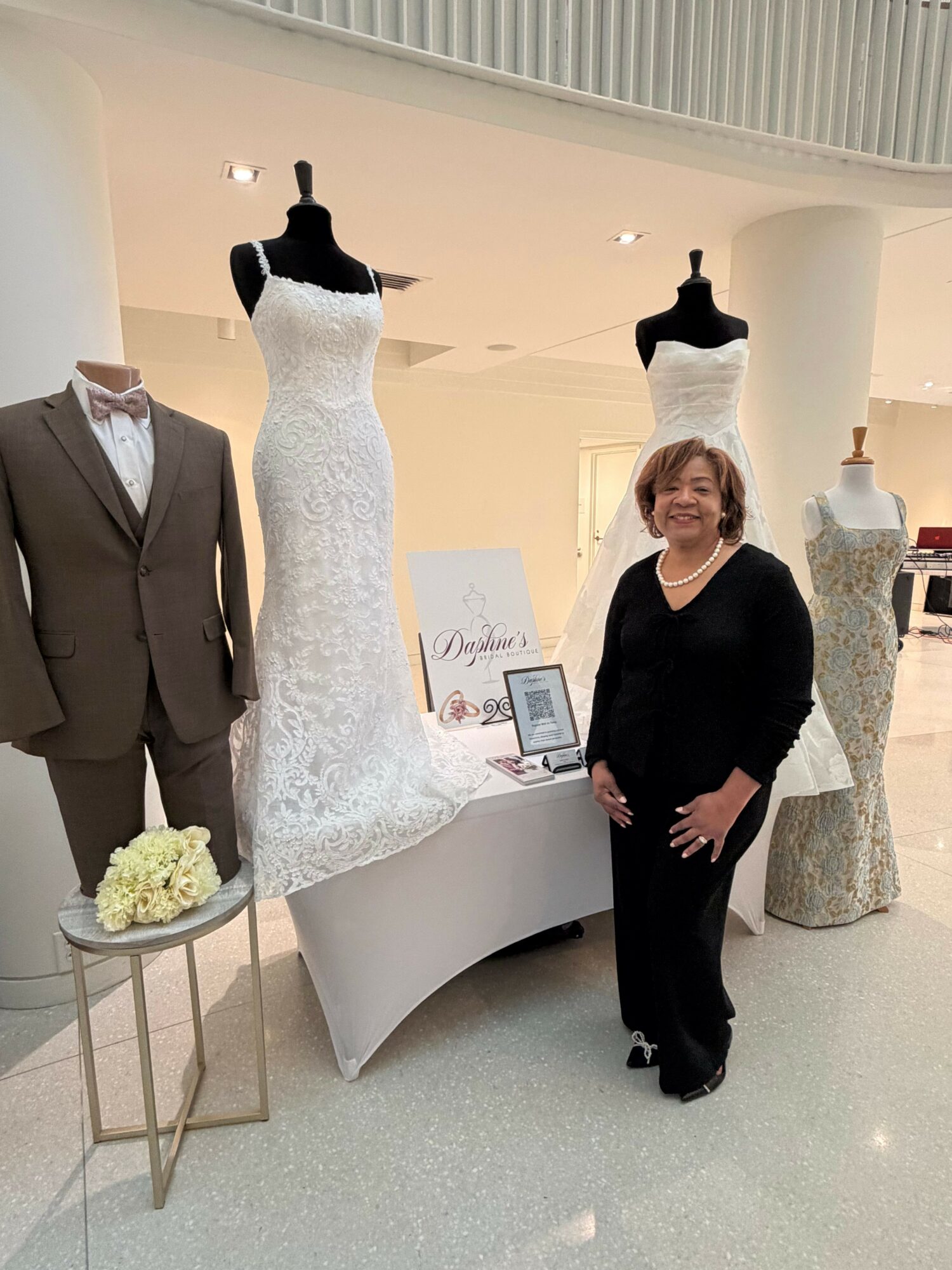 Woman in black dress standing next to a mannequin in a white dress, with a person in a suit holding flowers nearby.