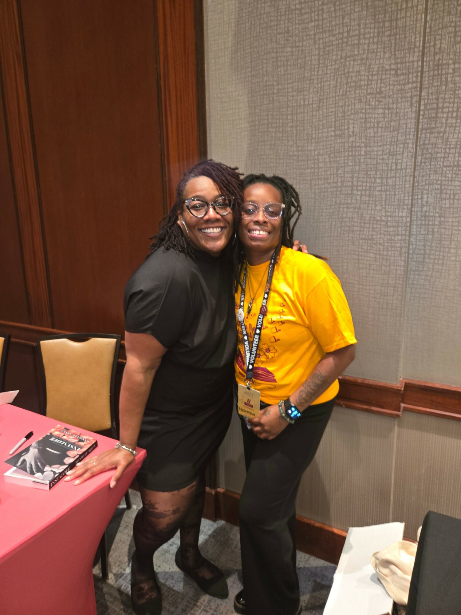 Two women standing close together, smiling, in a room with beige and wooden walls, one wearing a black outfit and the other a yellow shirt.