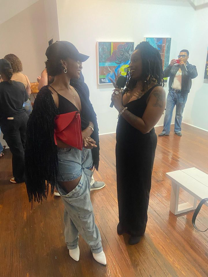 Two women with long hair and dark clothing converse in an art gallery, with paintings on the wall and a person taking a photo in the background.
