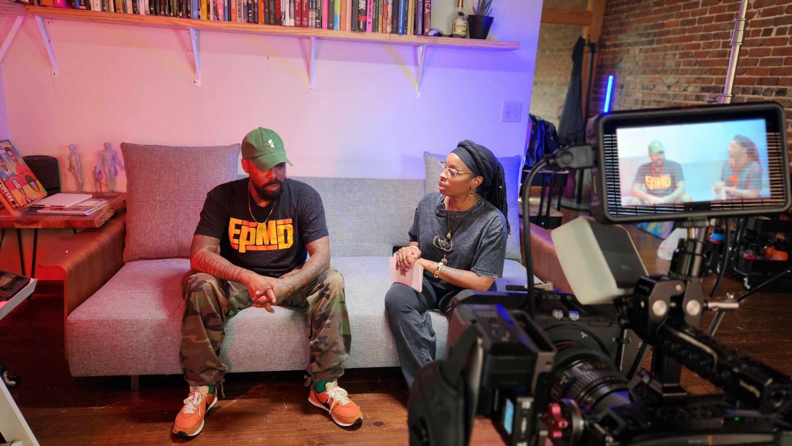 Two people sit on a sofa in a studio, being filmed with a camera, with a brick wall and shelves in the background.