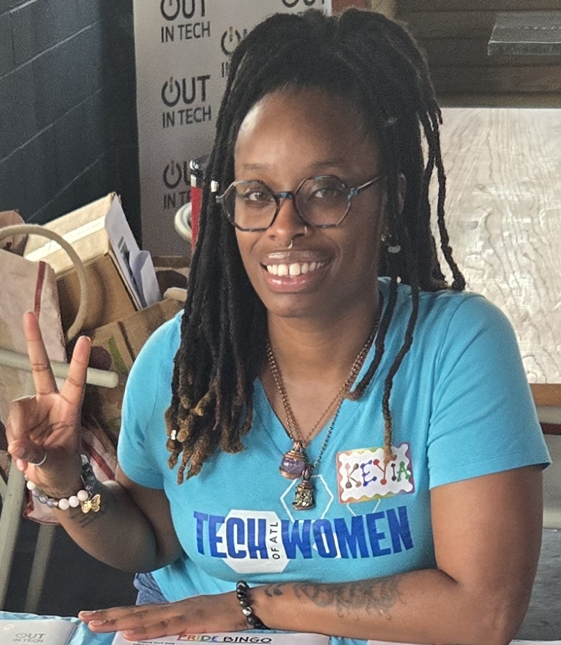Woman with glasses and dreadlocks smiling and making a peace sign, wearing a blue T-shirt with text, in a casual setting.