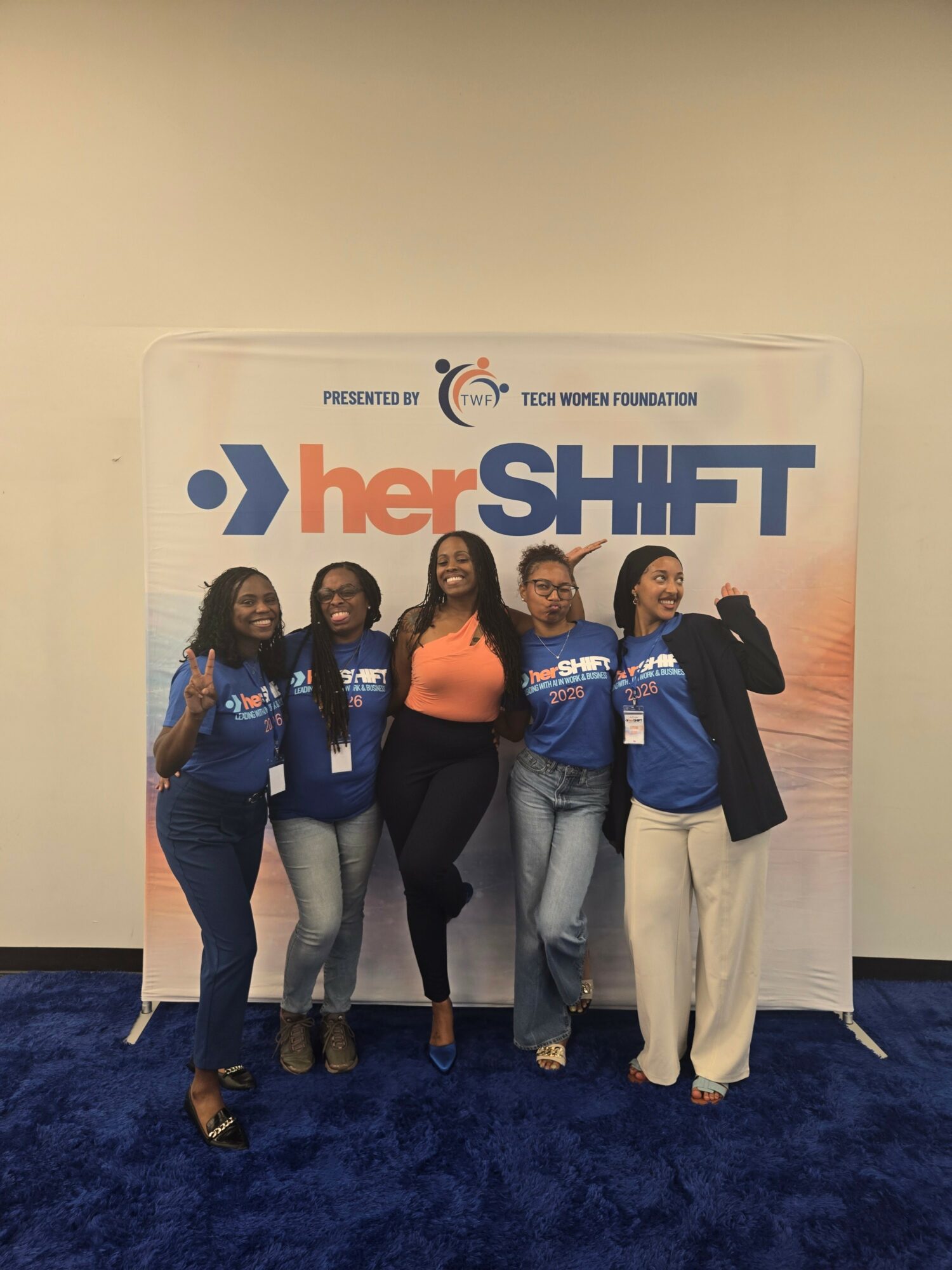Five women standing in front of a Her Shift banner, smiling and posing indoors. One woman is wearing an orange top.