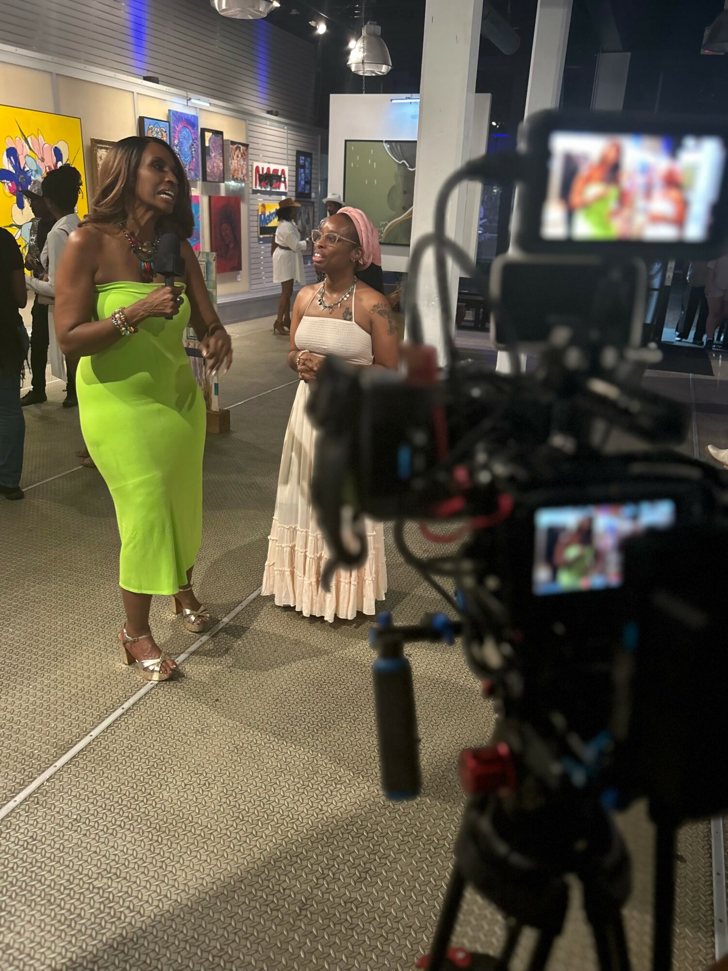Two women standing and talking in an indoor setting with display shelves and a camera in the foreground.