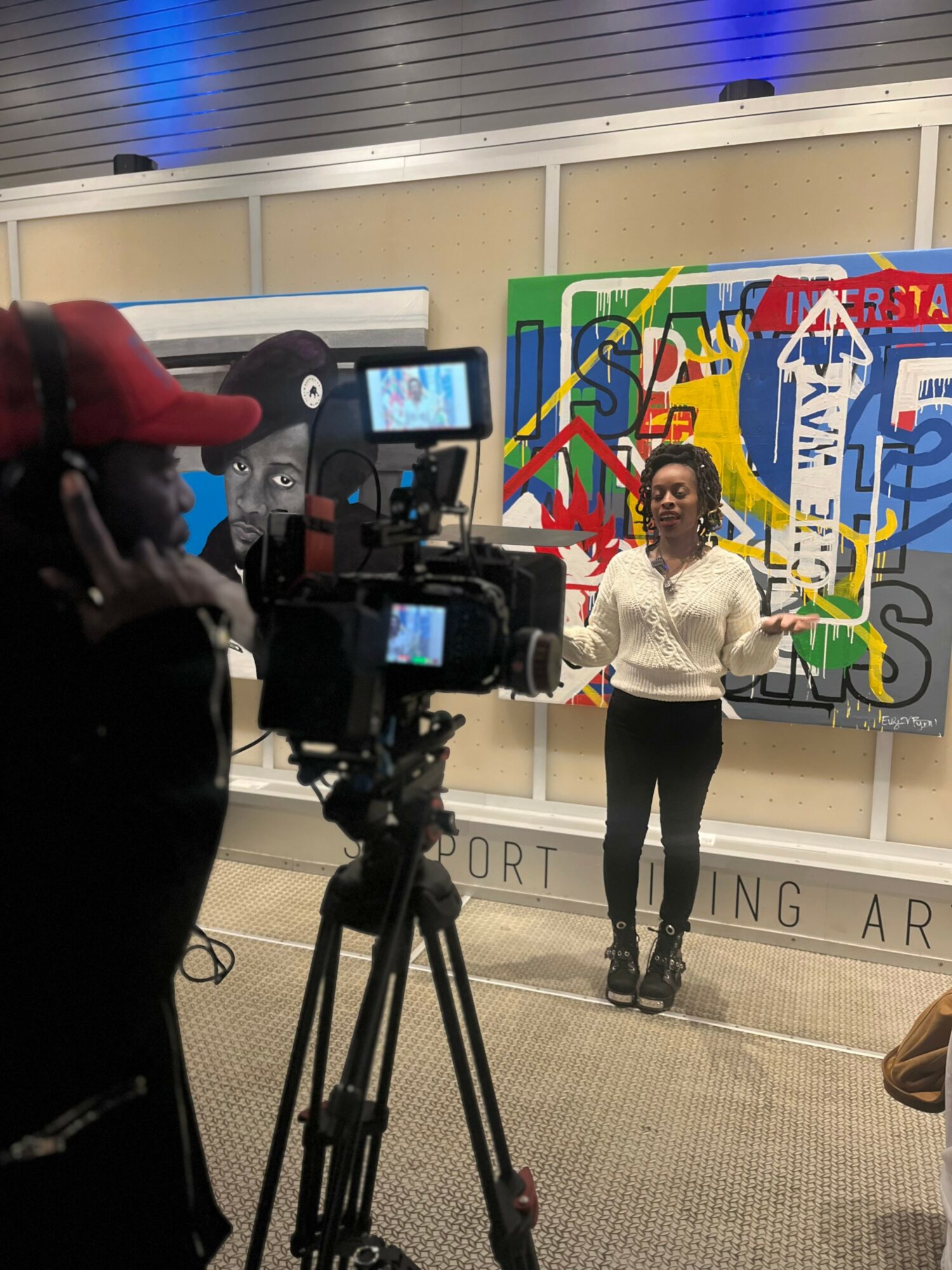 Woman standing in front of colorful abstract artwork, being filmed by a camera operator, in an indoor setting.