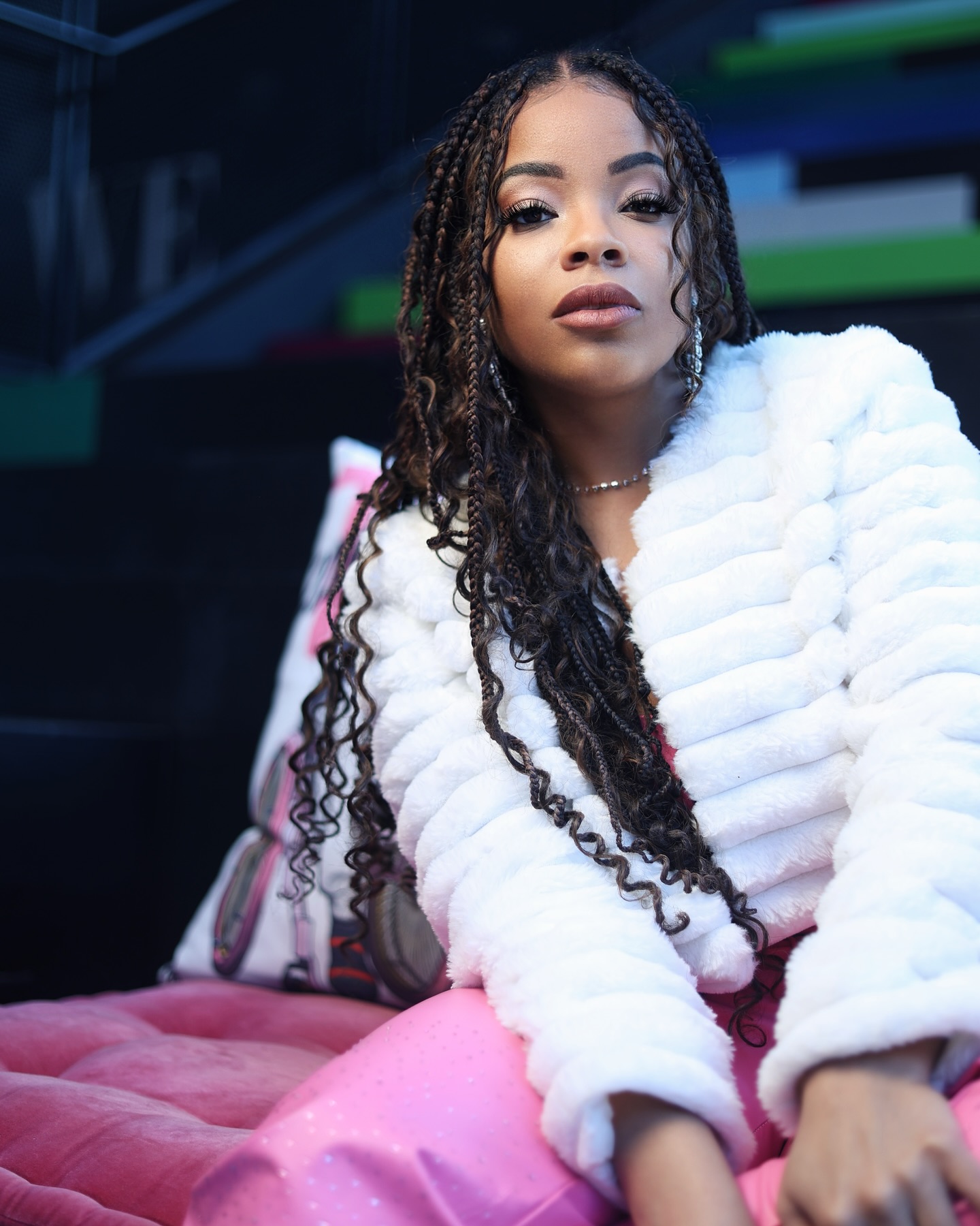 Young woman with long curly hair wearing a white fur coat and pink pants, sitting on a pink surface, looking at the camera.