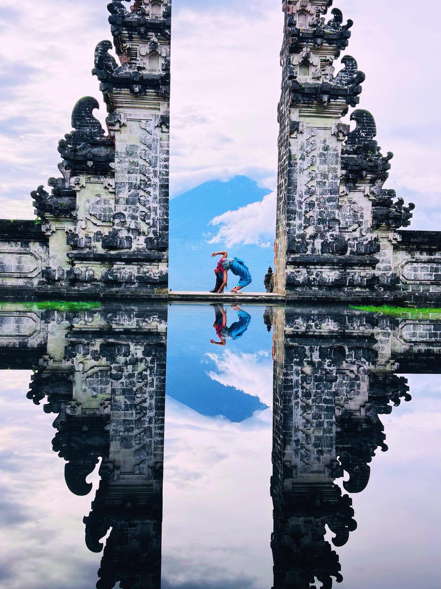 Person crouching between ornate stone gates with reflection in water, sky with clouds above, symmetrical scene.