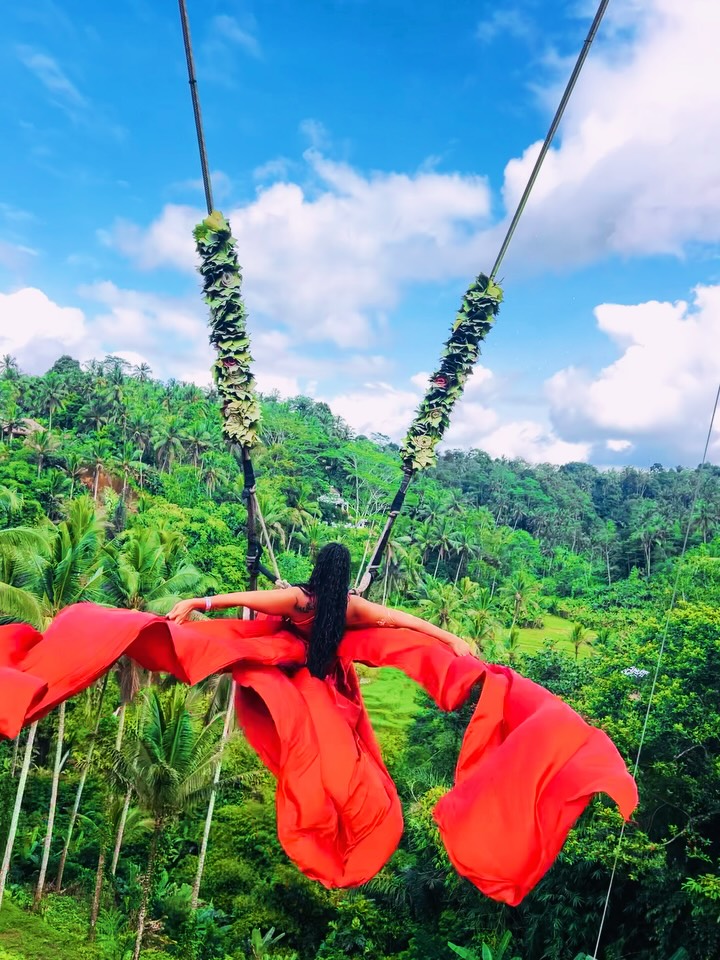 Person swinging on a red fabric swing with a lush green landscape and blue sky in the background.