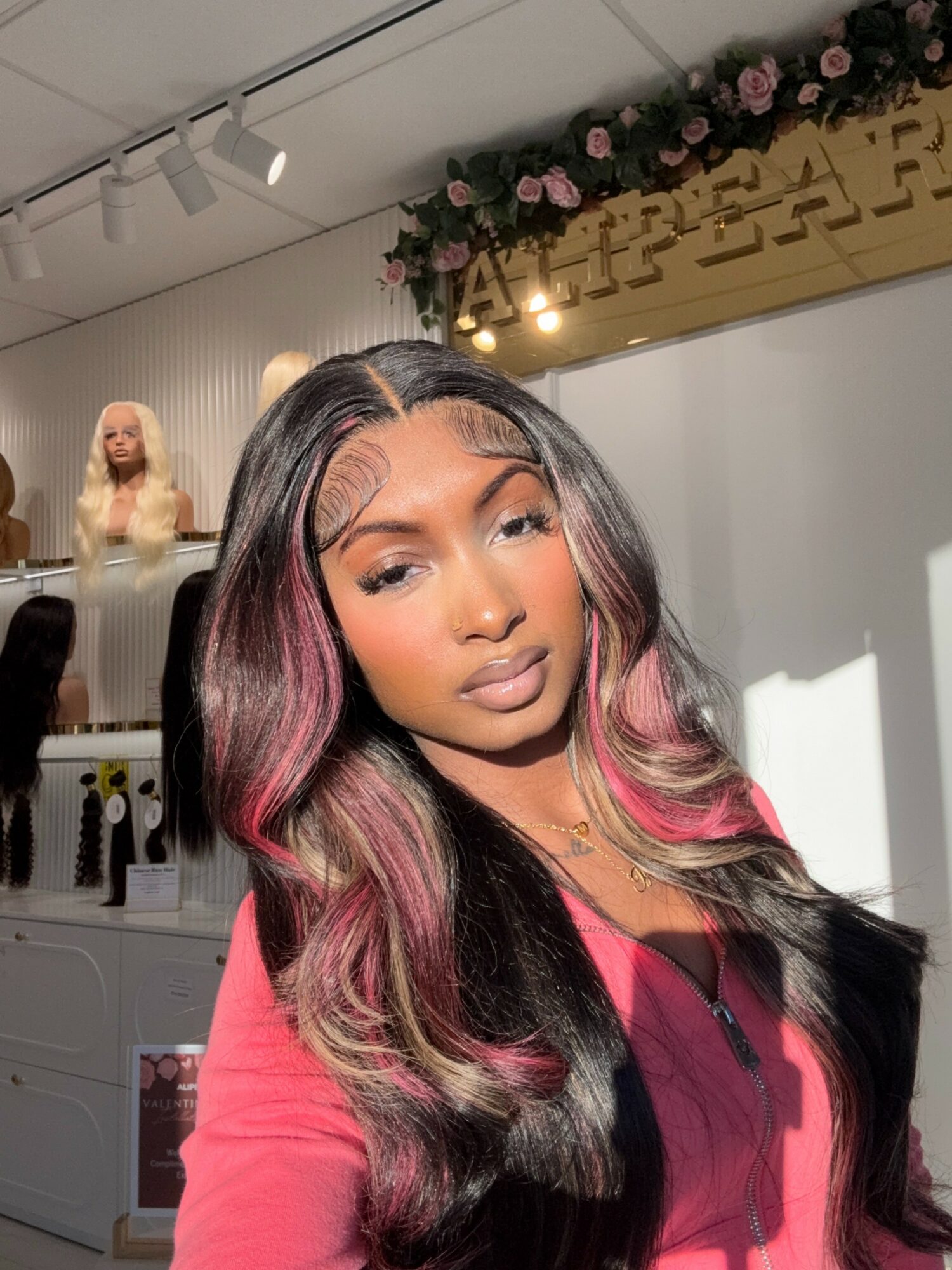 Young woman with long, wavy hair featuring pink highlights, posing indoors with a floral decoration and a sign in the background.