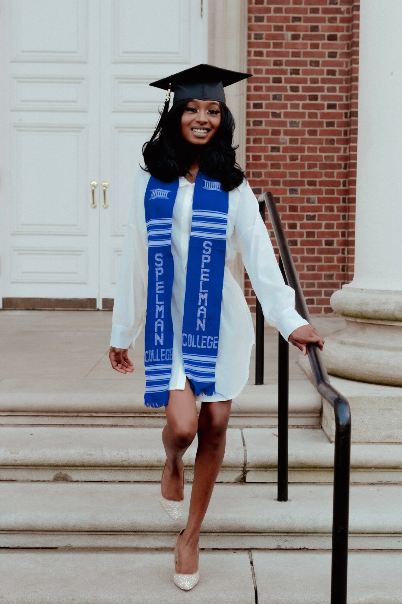 Young woman in graduation gown and cap, smiling, standing on steps outside building, holding a diploma, wearing a blue stole.