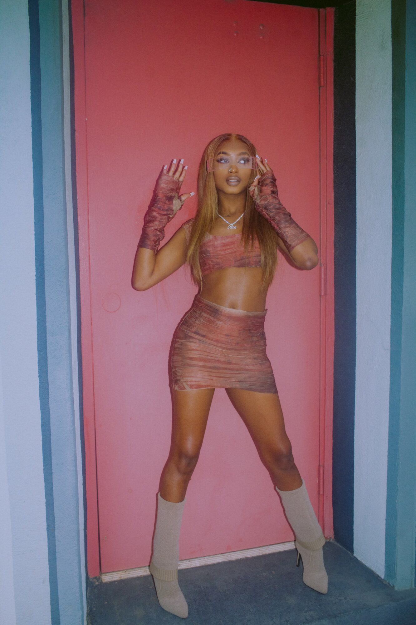 Young woman with long hair in a pink outfit standing in front of a pink door, hands near her face, looking at the camera.