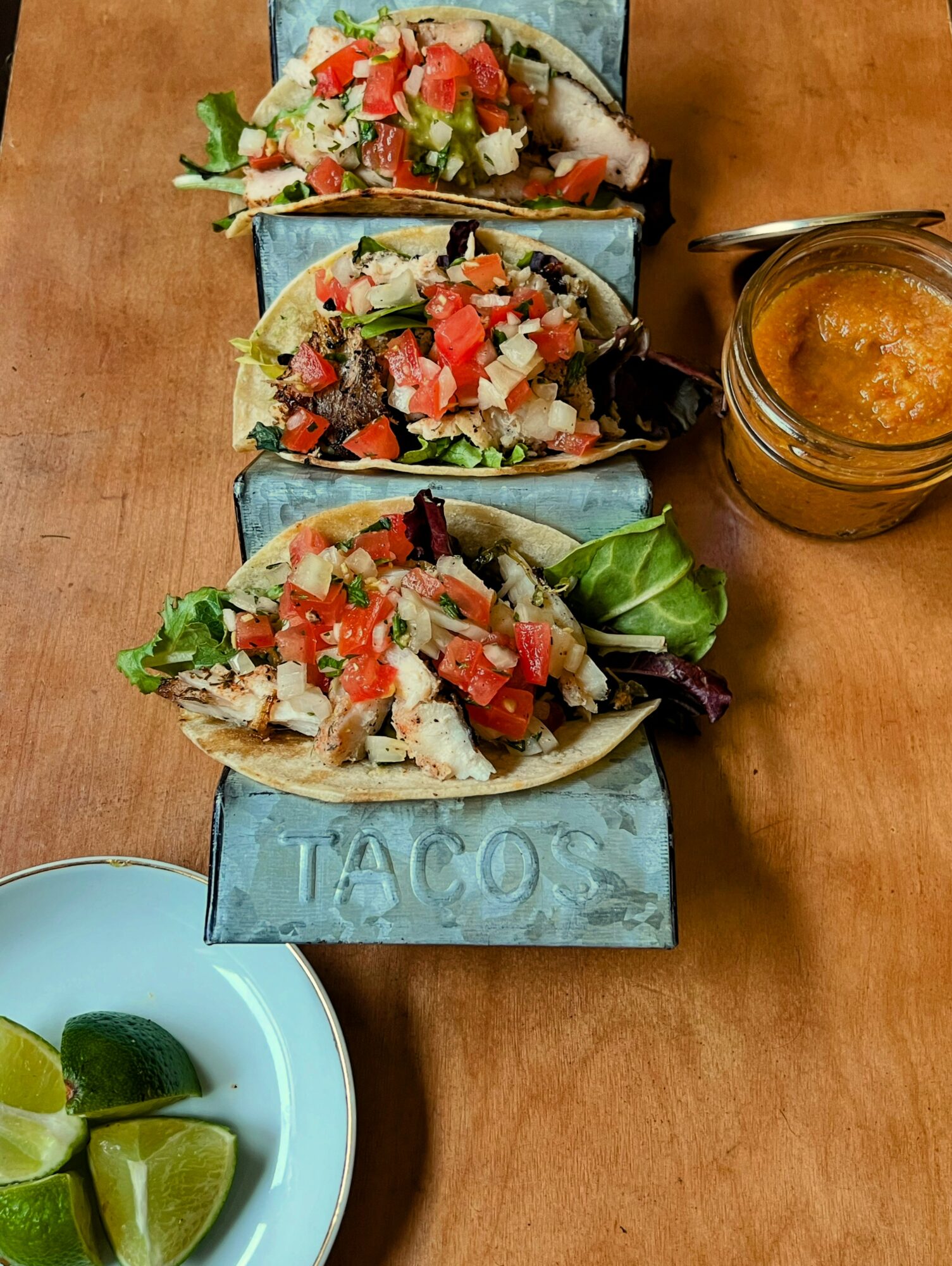 Three tacos with various fillings on a blue tray, a jar of sauce, and a bowl of lime wedges on a wooden surface.