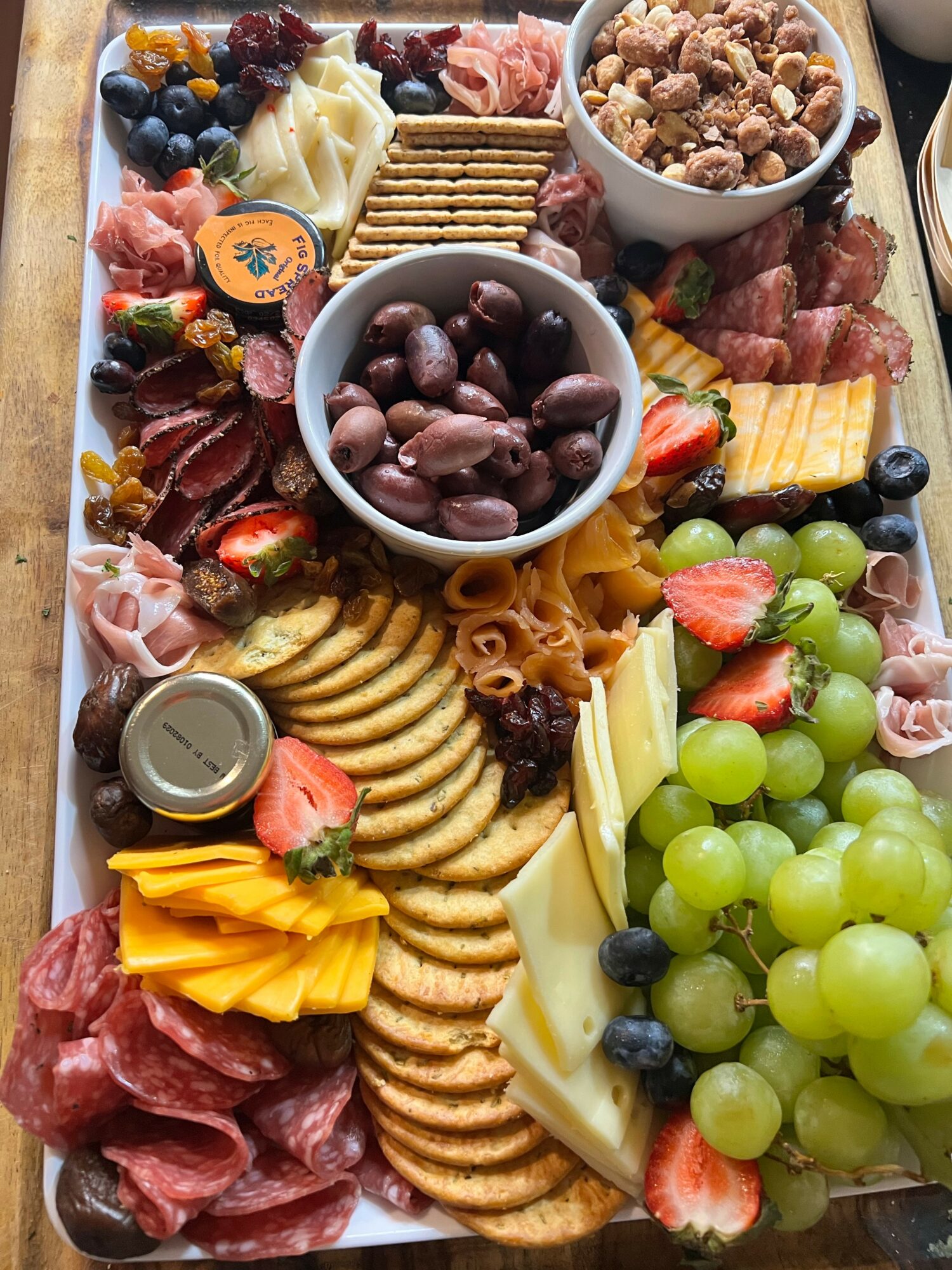 Assorted fruits, cheeses, crackers, nuts, and cured meats arranged on a platter with two bowls of grapes and nuts.