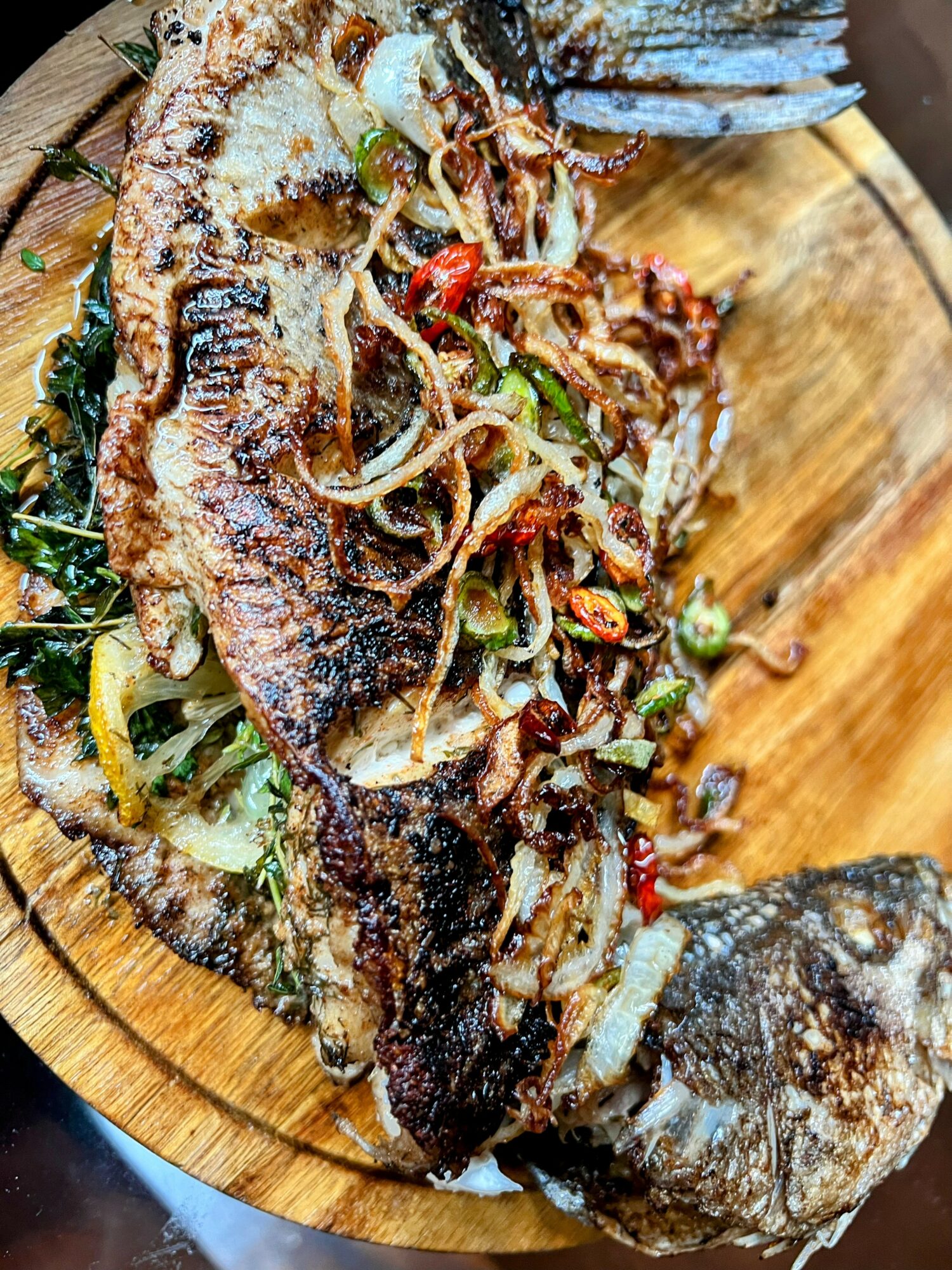 Grilled fish on a wooden platter with onions, herbs, and vegetables, garnished with herbs and spices.
