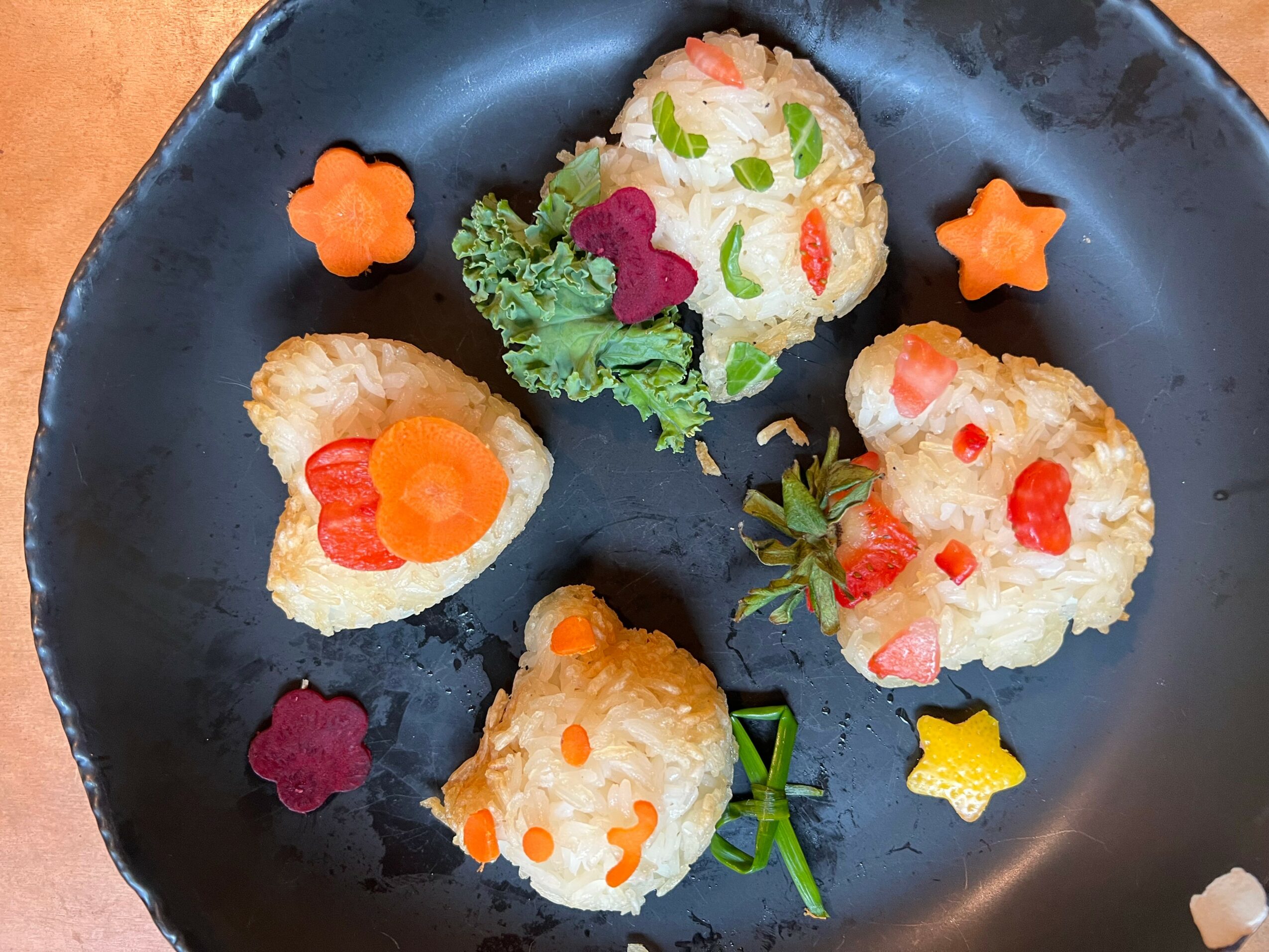 Plate with rice balls decorated with vegetables, garnished with vegetables and flower-shaped decorations, on a black plate.