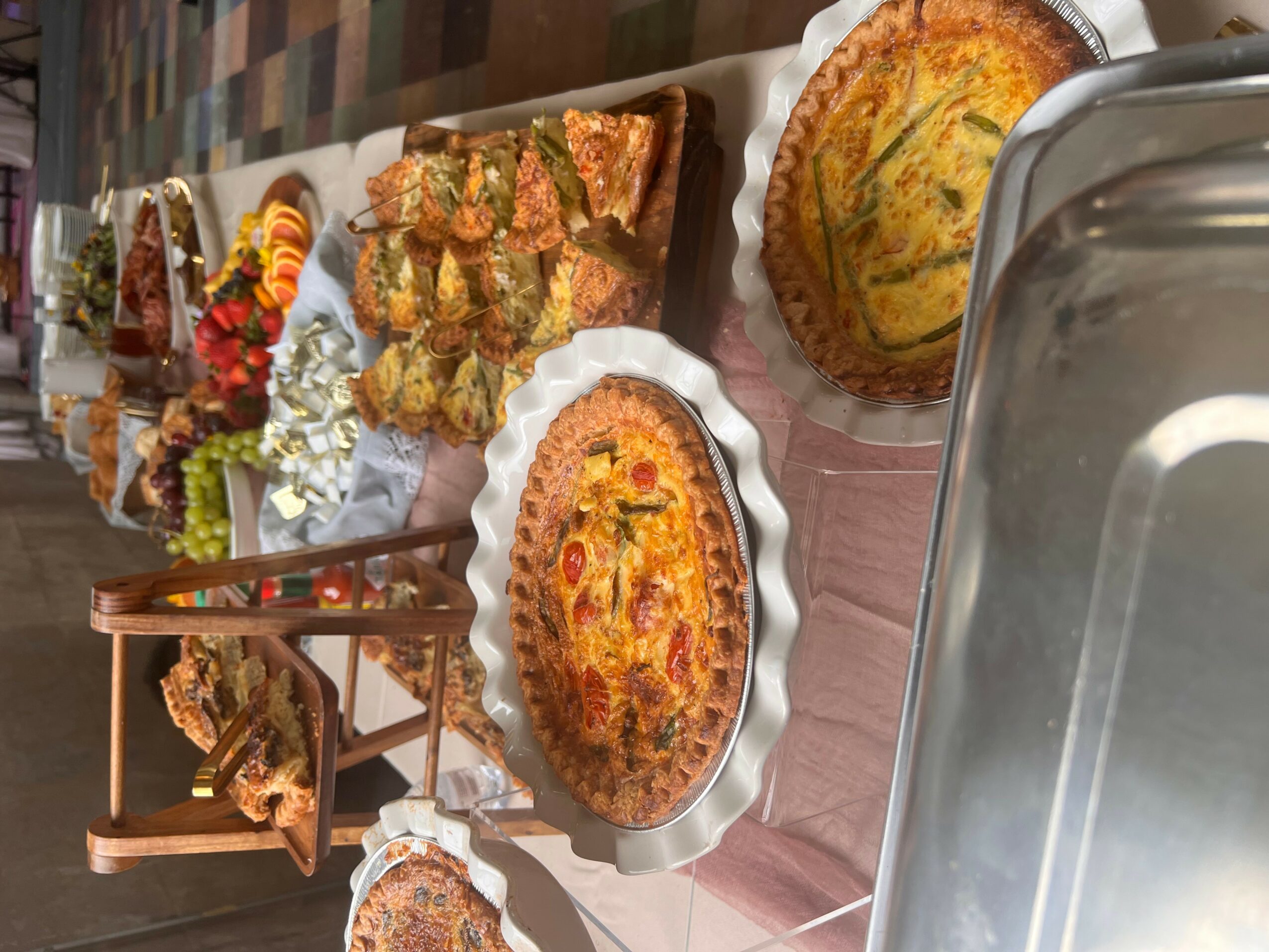 Table with various baked goods, including pies and cakes, displayed on plates and stands, with flowers in the background.
