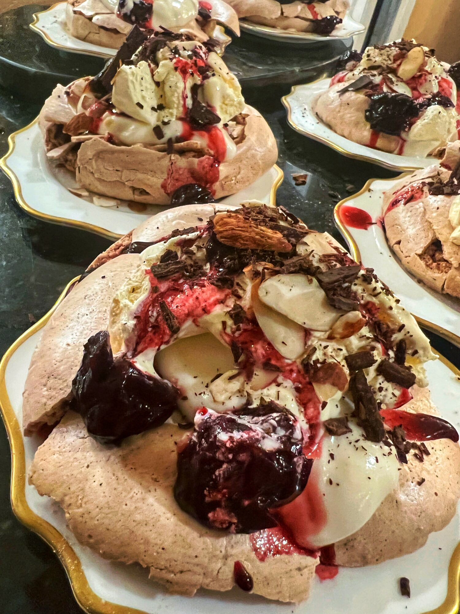 Multiple plates with large, decorated desserts topped with various chocolates, fruits, and sauces, on a dark surface.