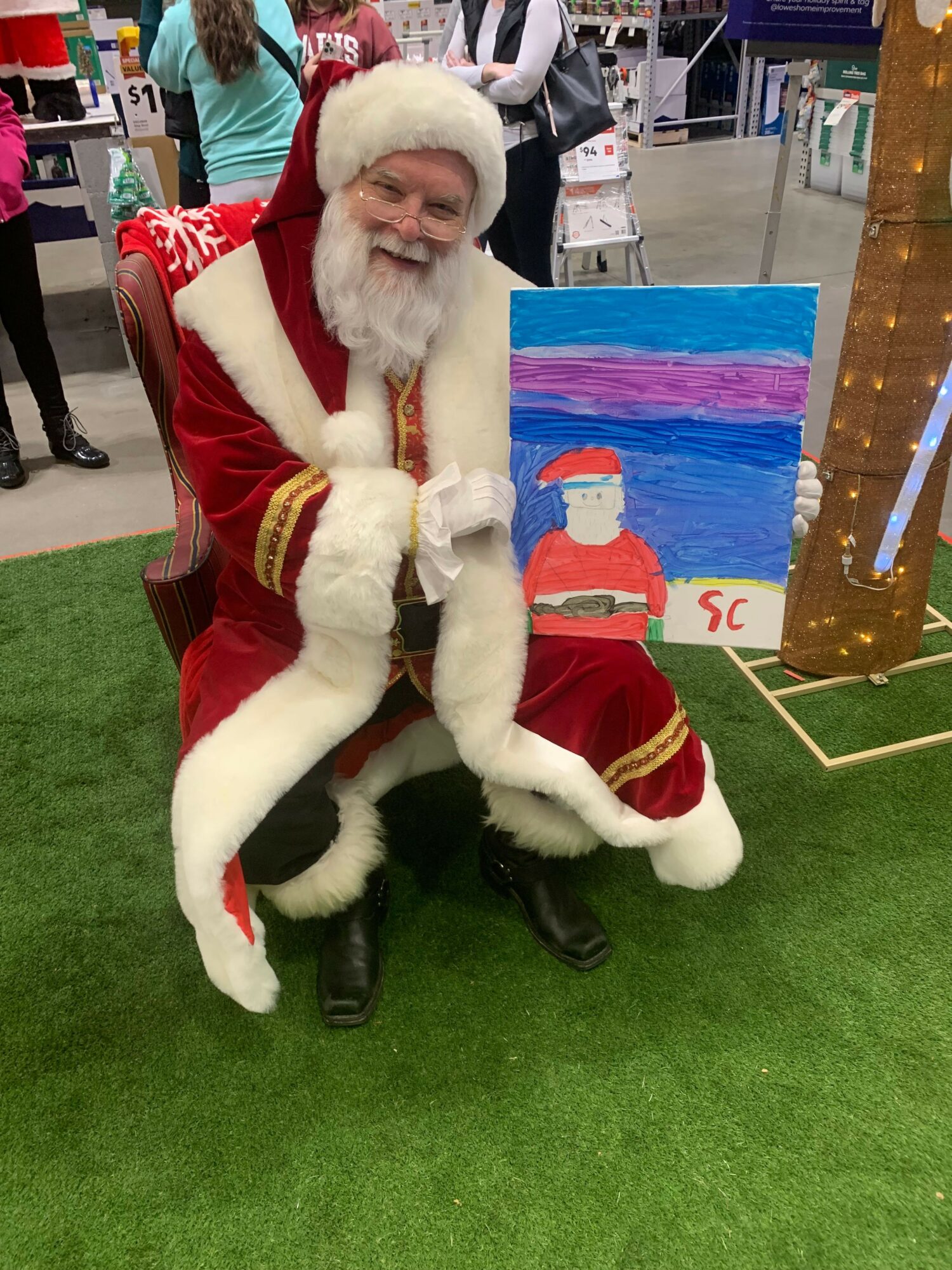 Person dressed as Santa Claus sitting on green artificial grass, smiling, with a colorful painting in the background.