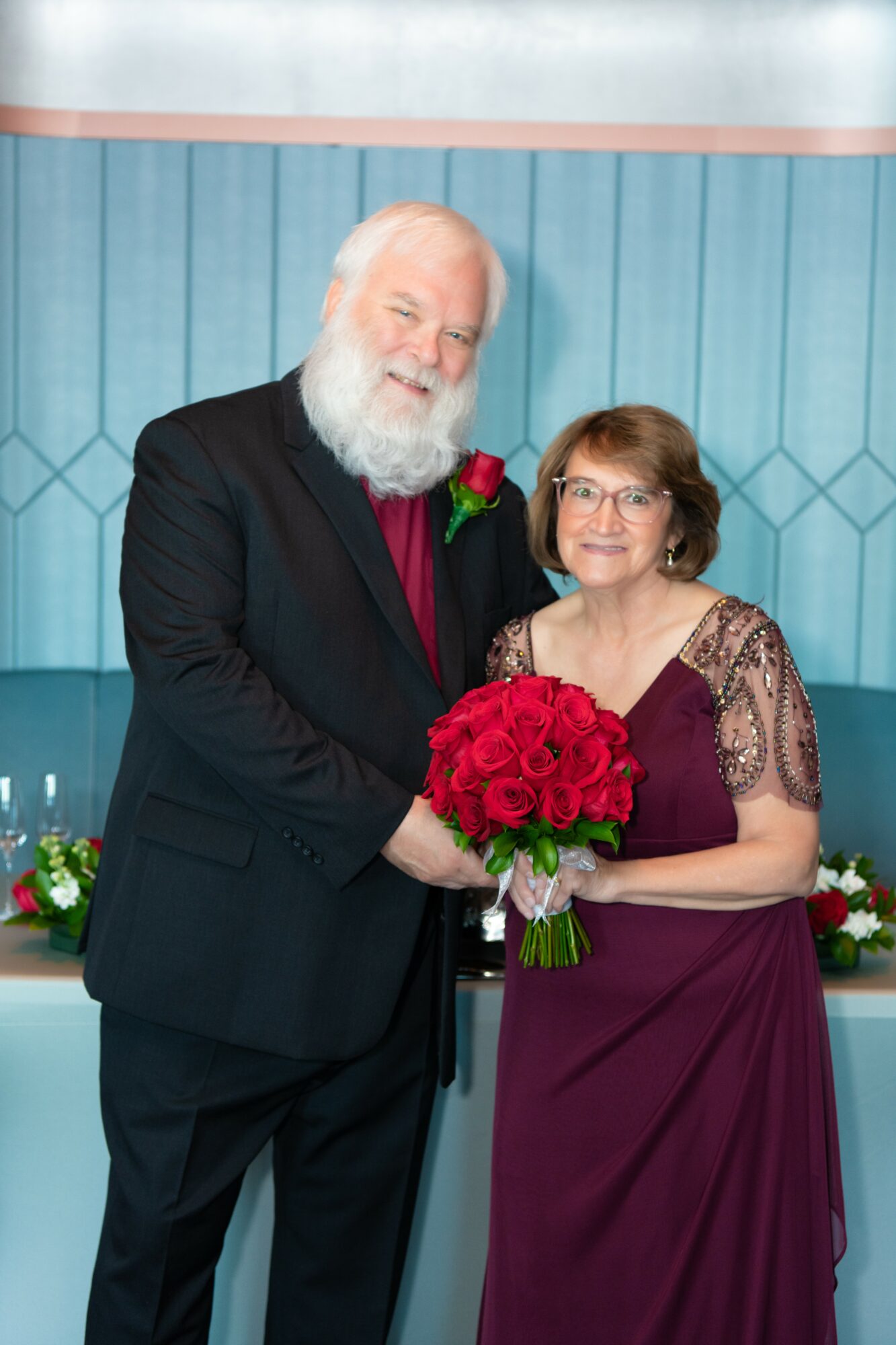 An elderly man with a white beard and a woman with glasses holding a bouquet of red roses, smiling together.