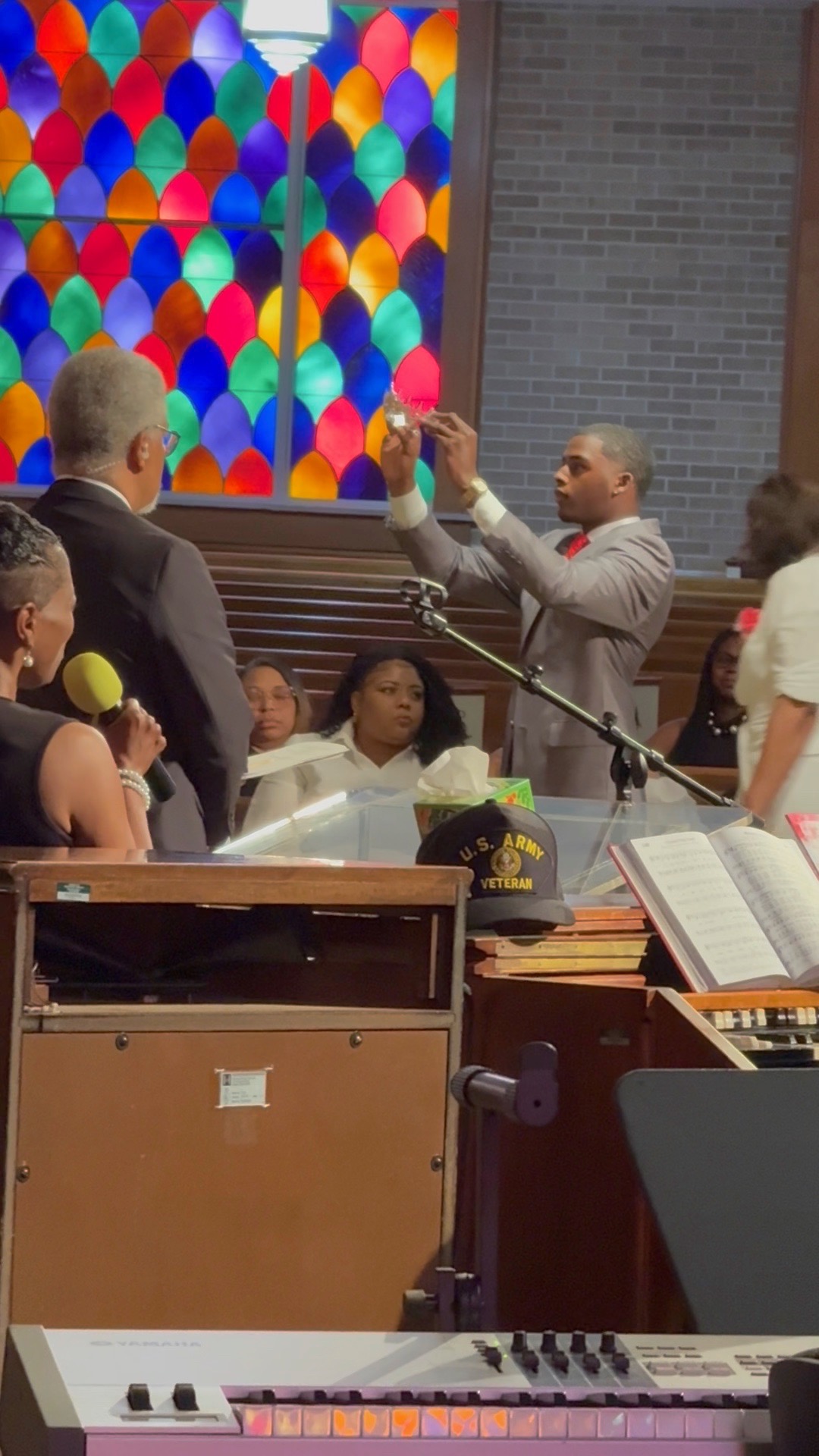 Man conducting choir with children and adults in church, colorful stained glass window in background.