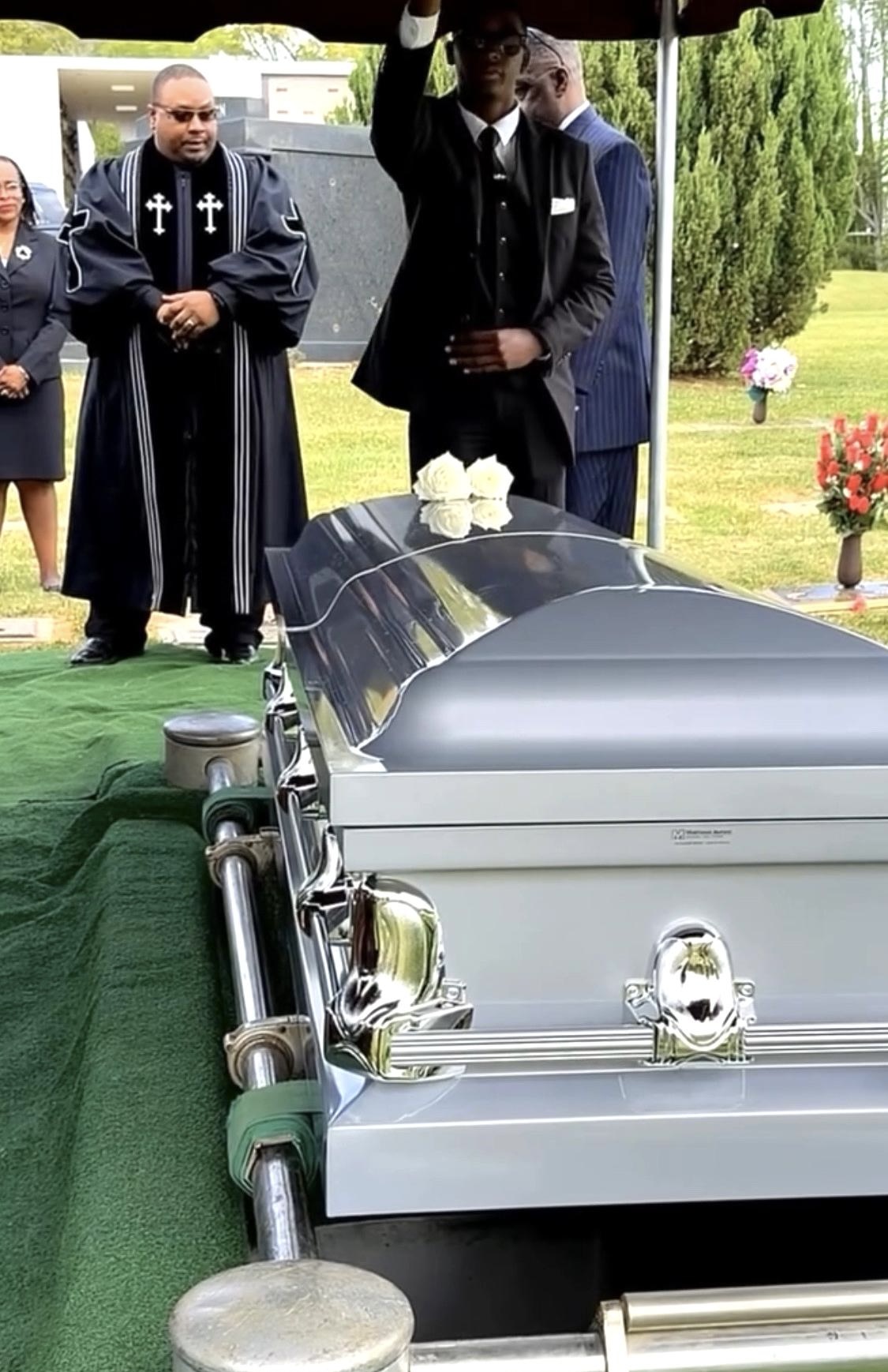 People gathered at a funeral with a silver casket, some wearing black, outdoors with greenery and flowers present.