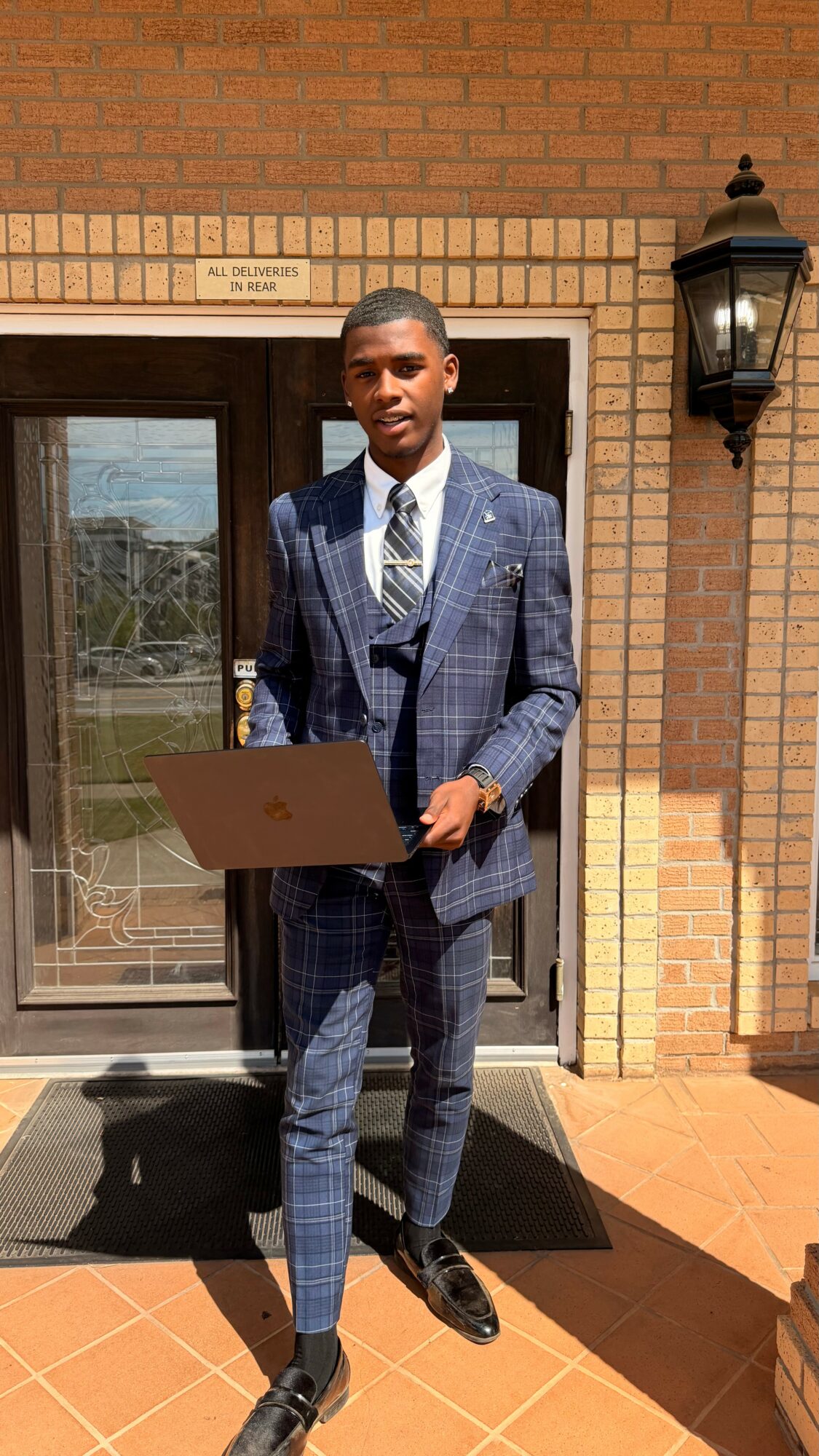 Young man in a blue checkered suit holding a laptop, standing outside a brick building with a glass door and lantern.
