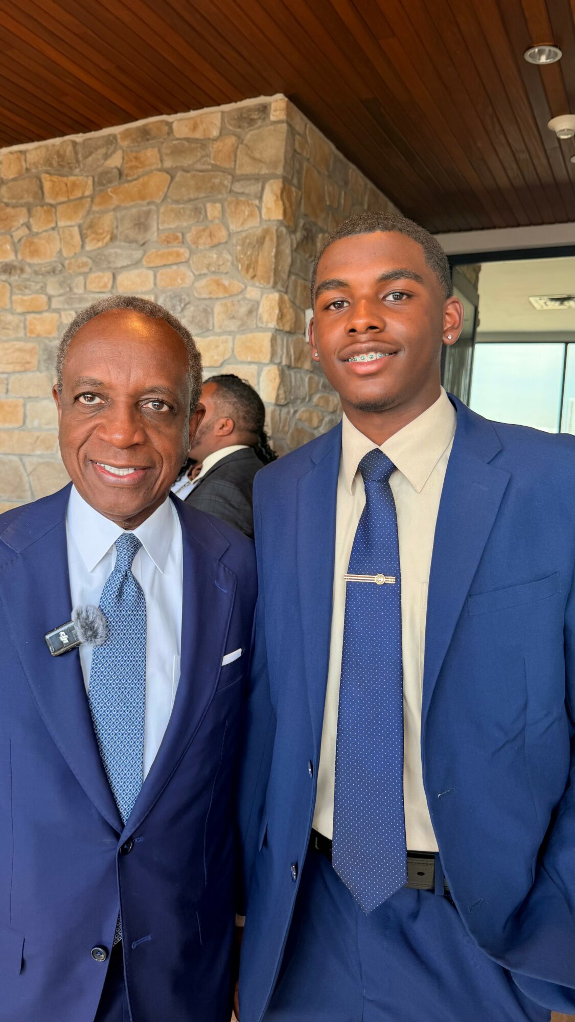 Two men in suits smiling, standing indoors near a stone wall and wooden ceiling.