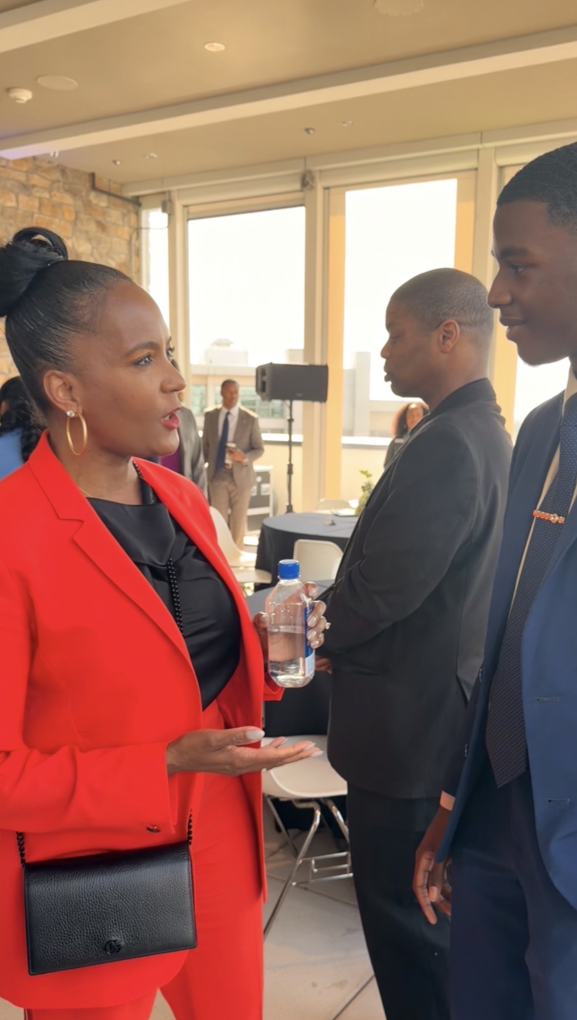 Three people in business attire engaged in conversation indoors, with large windows in the background.