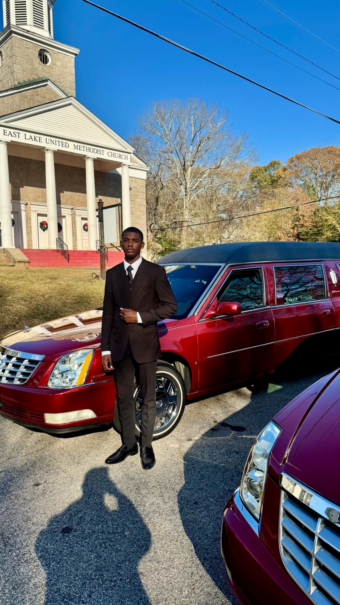 Man in black suit standing next to a red limousine in a parking lot, with a church building in the background.
