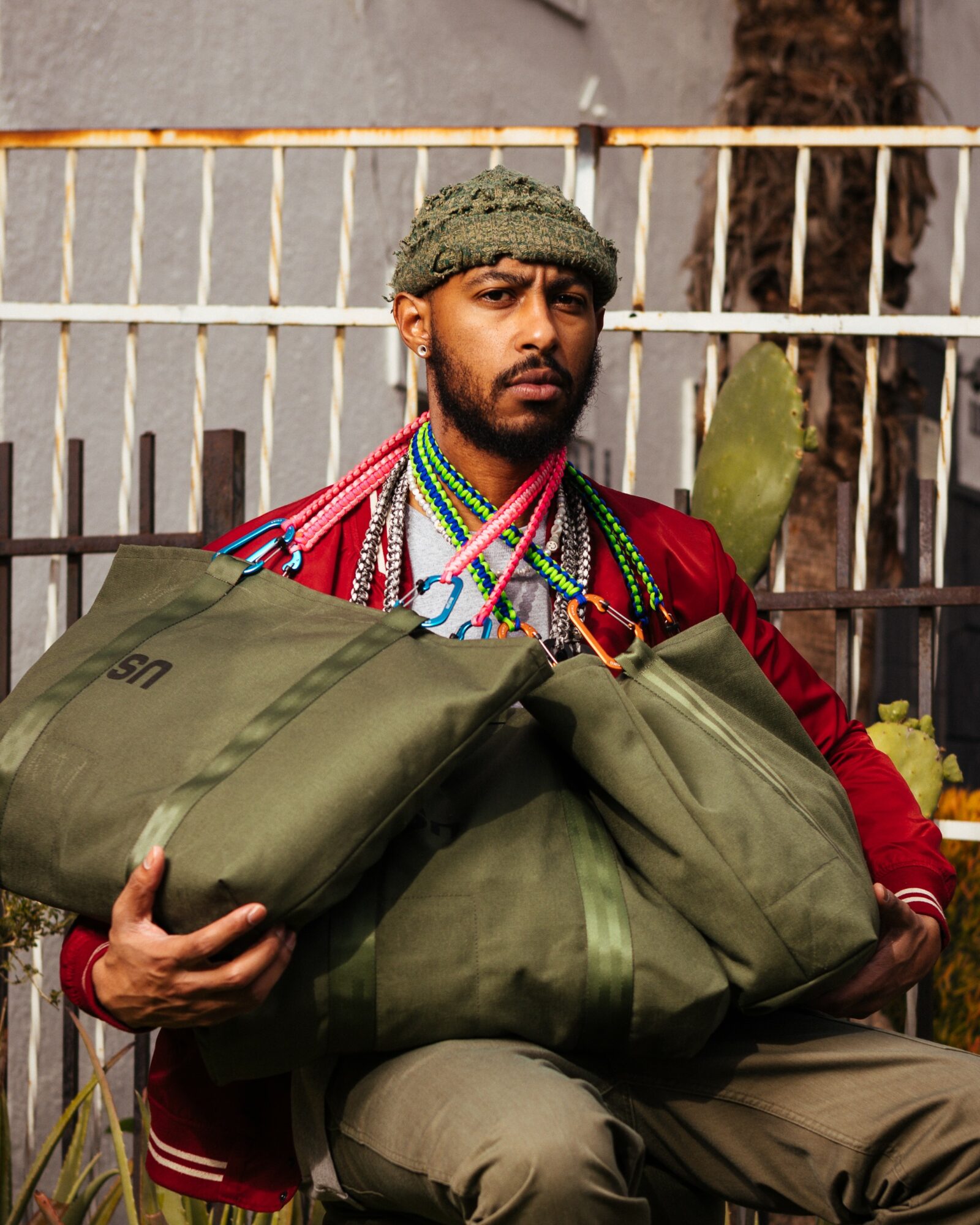 Man with a beard and knit cap holding large green bags outdoors, fence and plants in background.