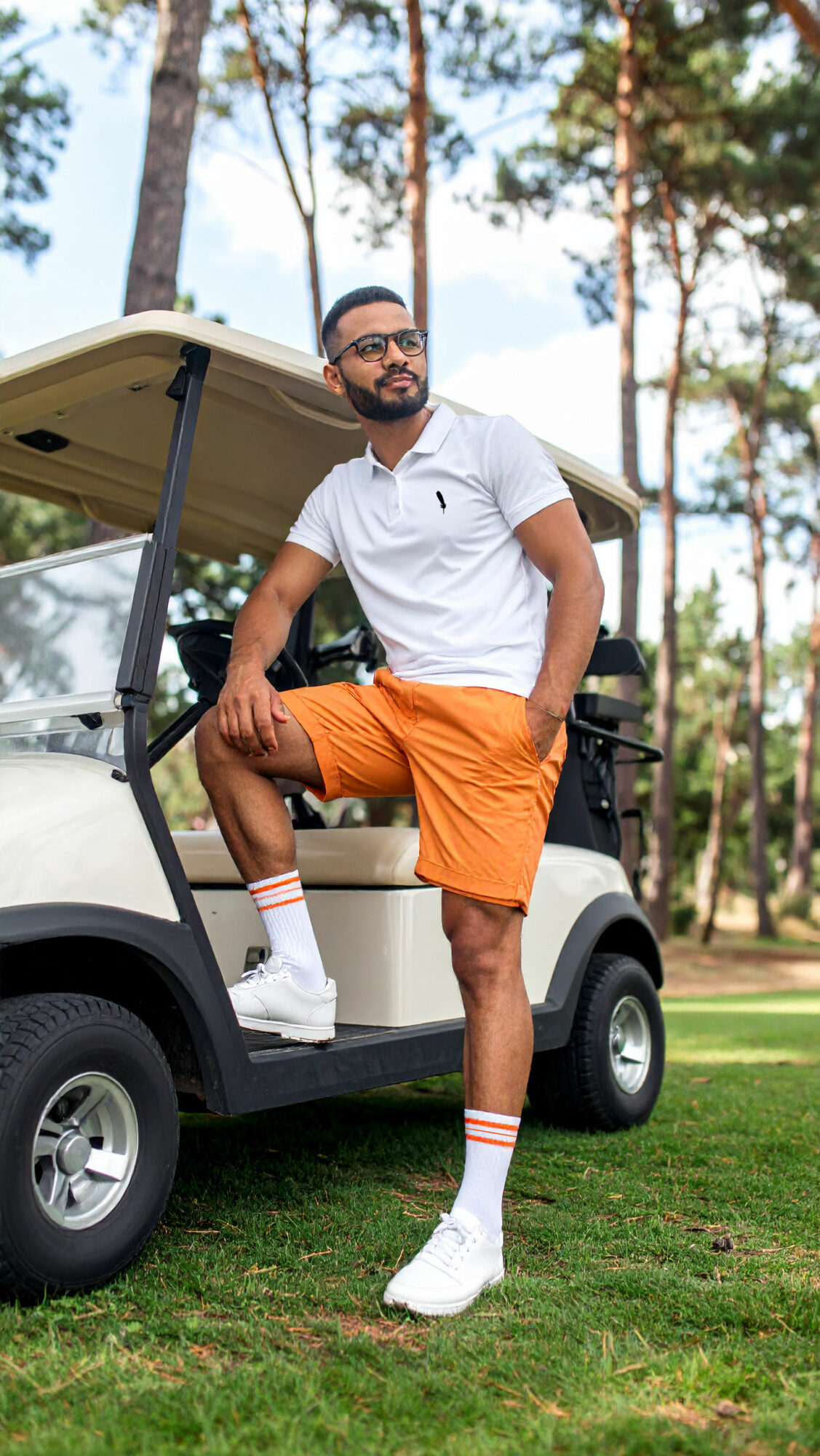Man leaning on golf cart in a park with trees, wearing white polo shirt, orange shorts, and white sneakers.