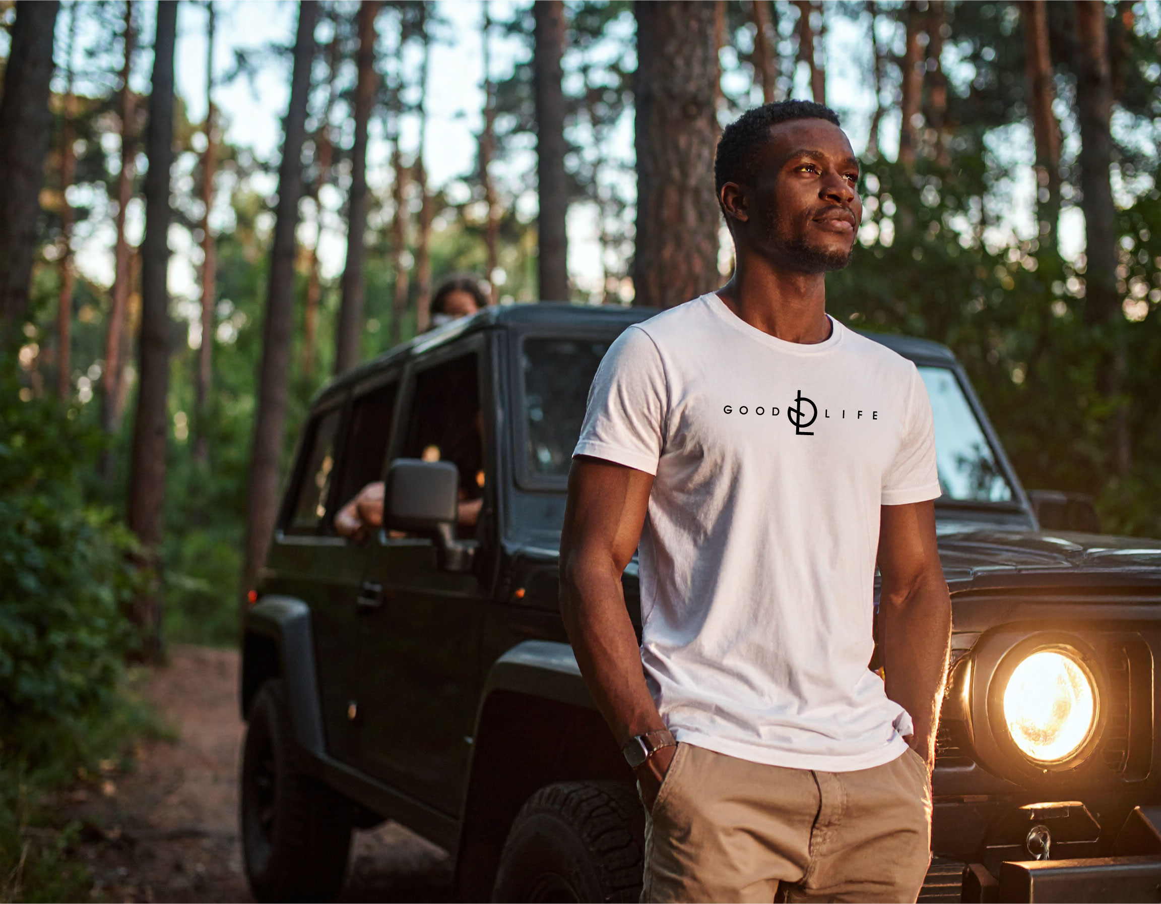 Man standing outdoors near a black vehicle in a forest, wearing a white t-shirt and beige pants.