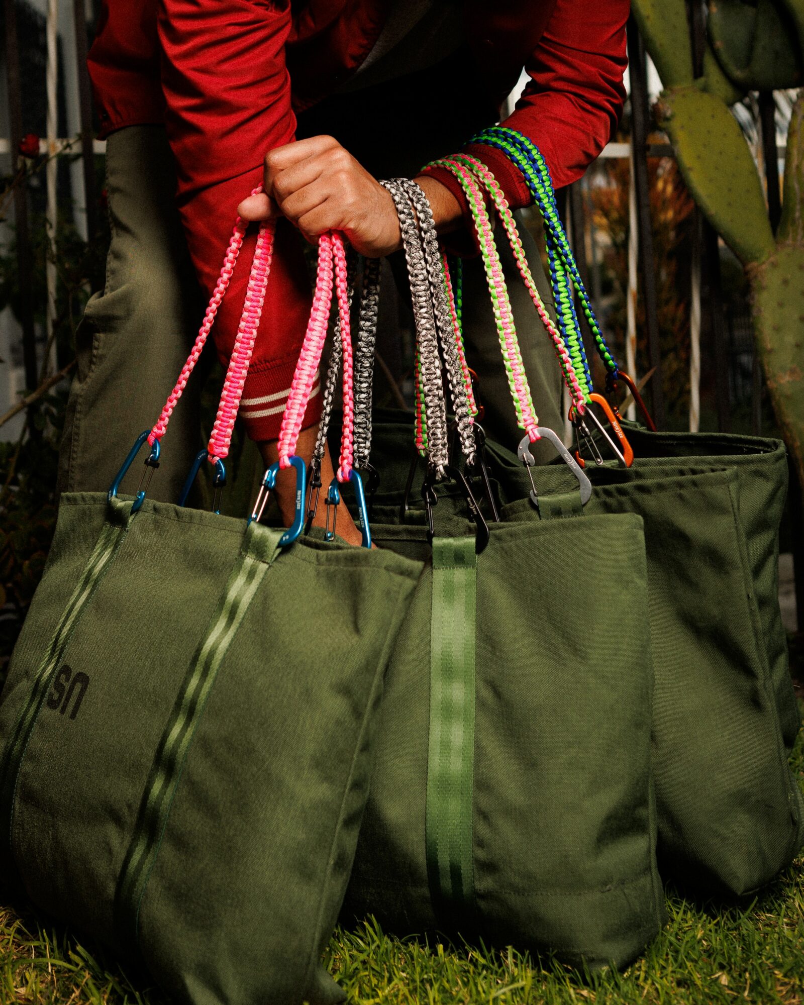 Person holding multiple colorful beaded bags with green bags on the ground, outdoors, with plants in the background.