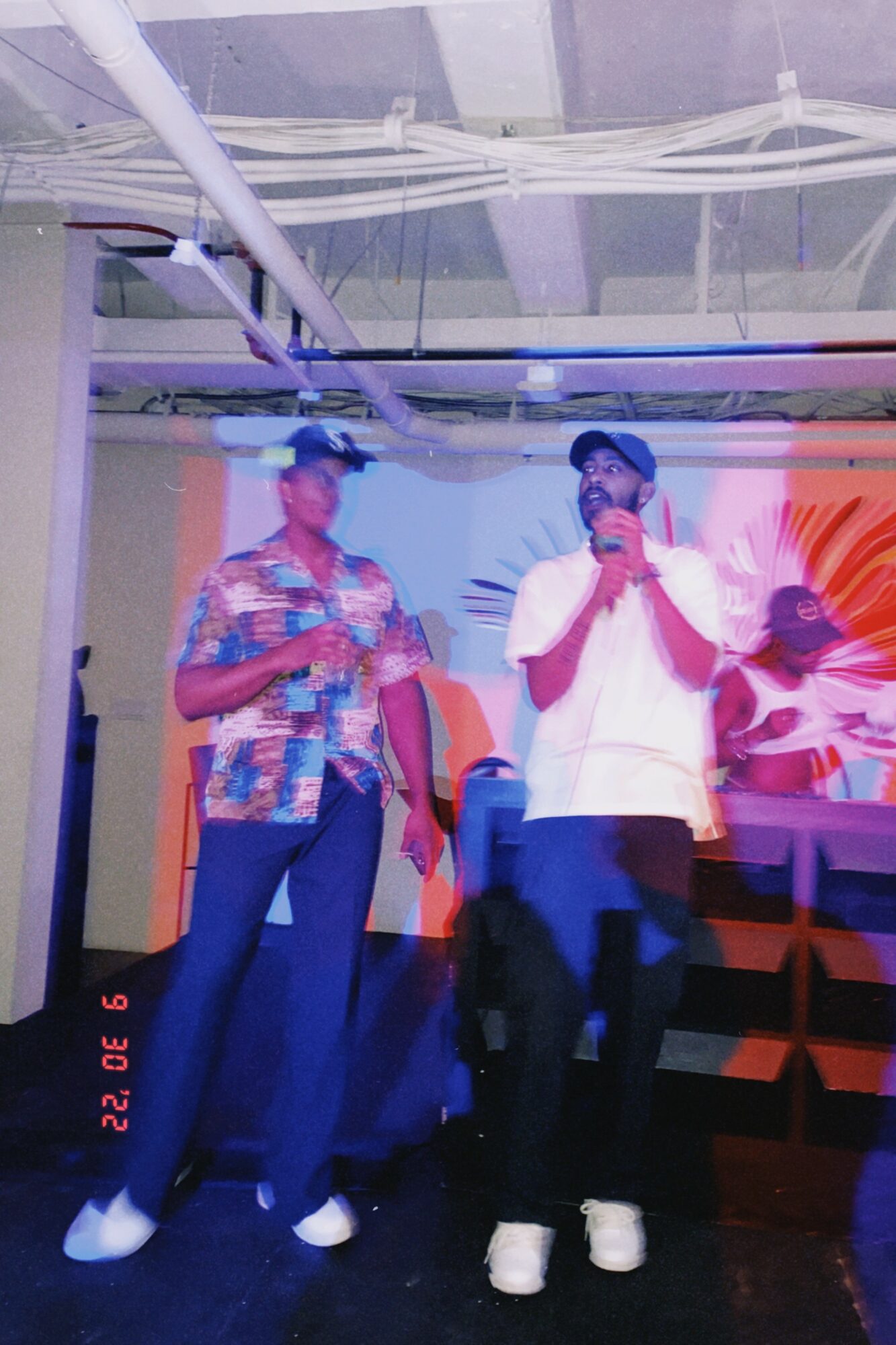 Two people stand on stage with a colorful background, one speaking into a microphone, the other holding a phone.