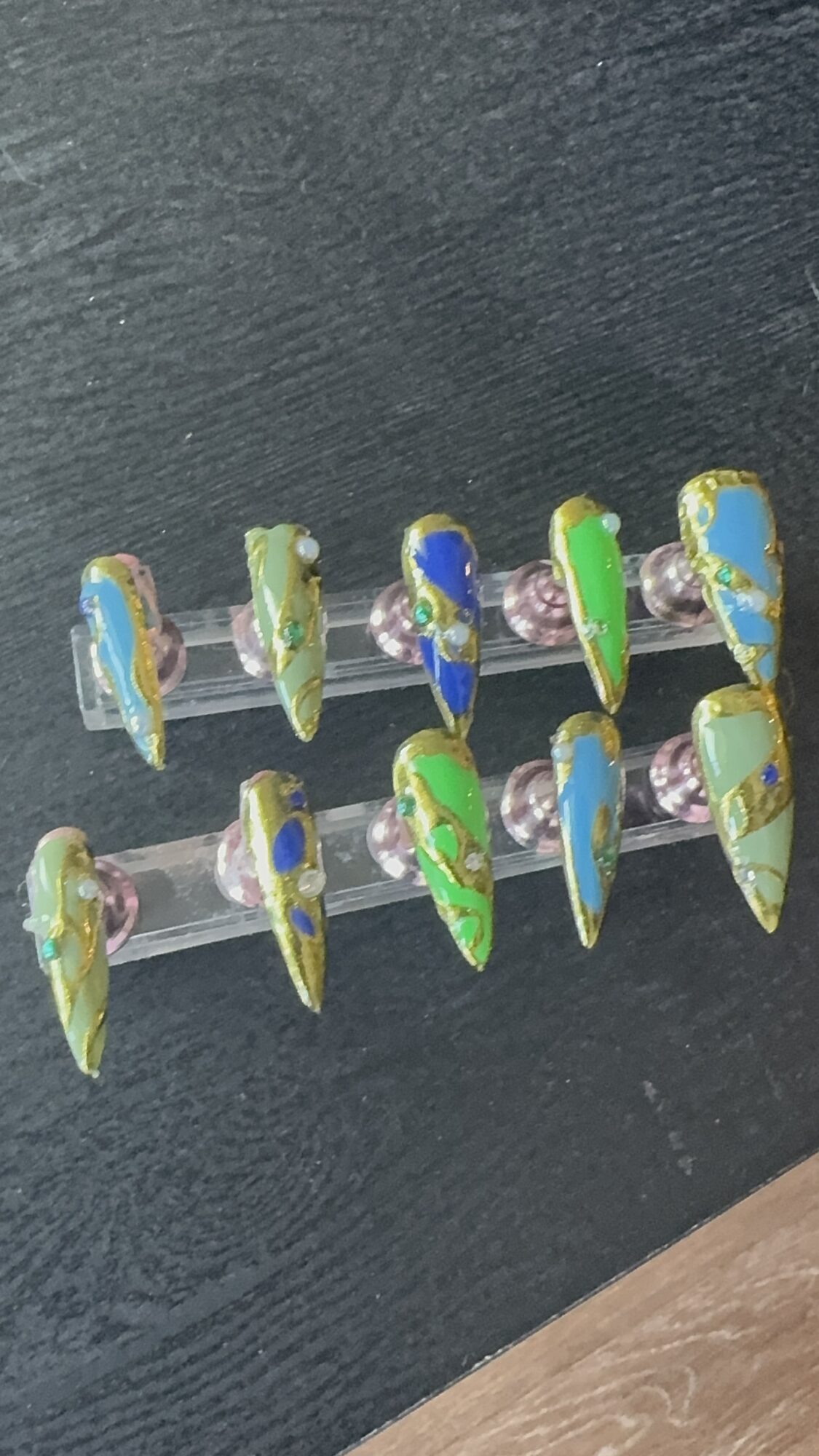Nail art with colorful, abstract designs on artificial nails arranged on a display stick, against a dark background.