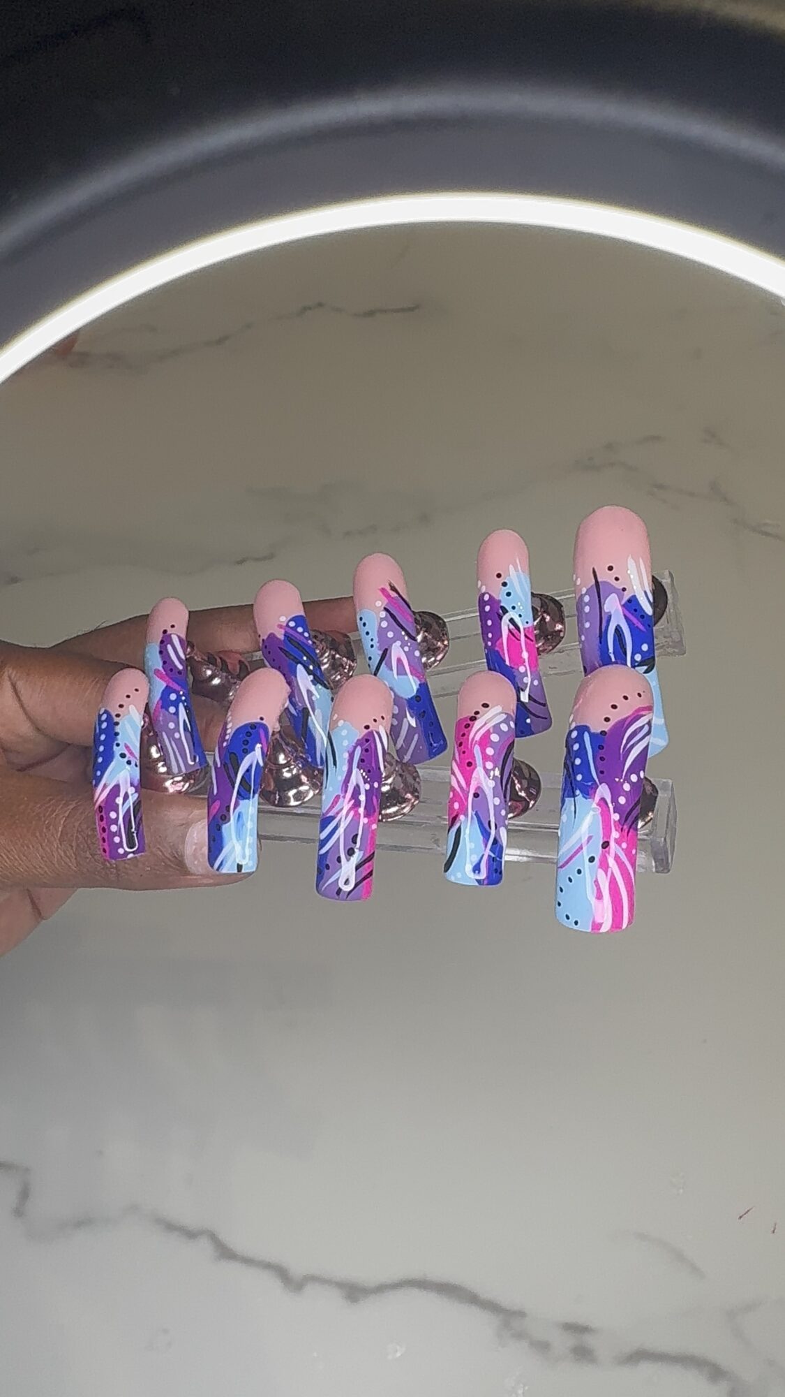 Hand holding long, decorated nails with blue, pink, and purple designs, including waves and abstract patterns, against a marble surface.