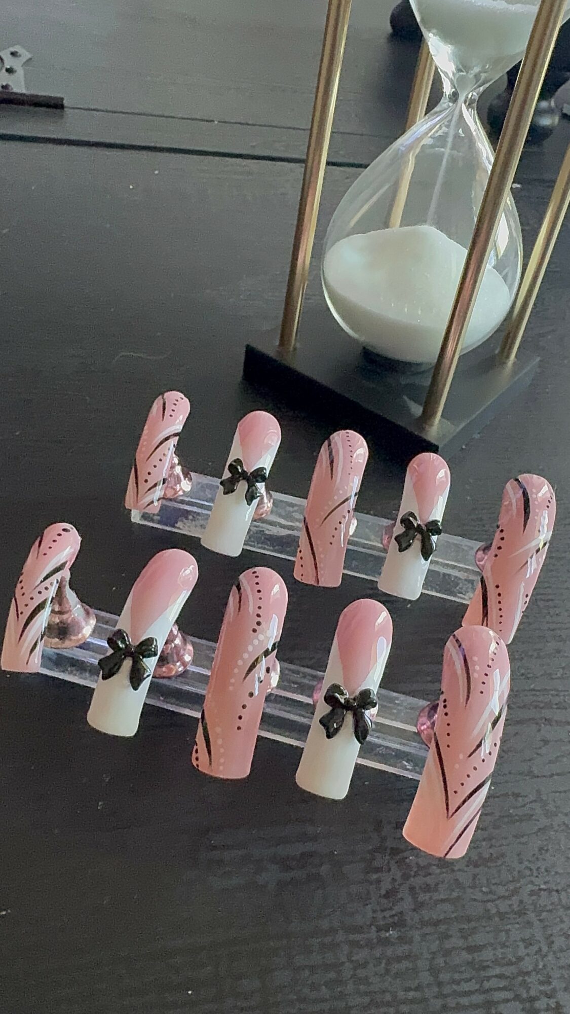 Decorative pink and white nails with black bows, arranged on a dark surface near an hourglass and gold frame.