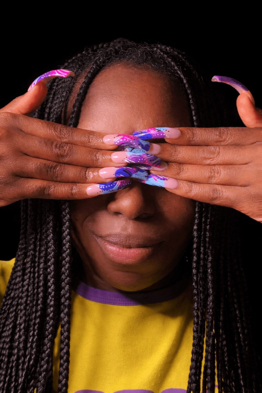 Person with long braided hair covering eyes with hands, colorful long nails, smiling, wearing yellow and purple shirt.