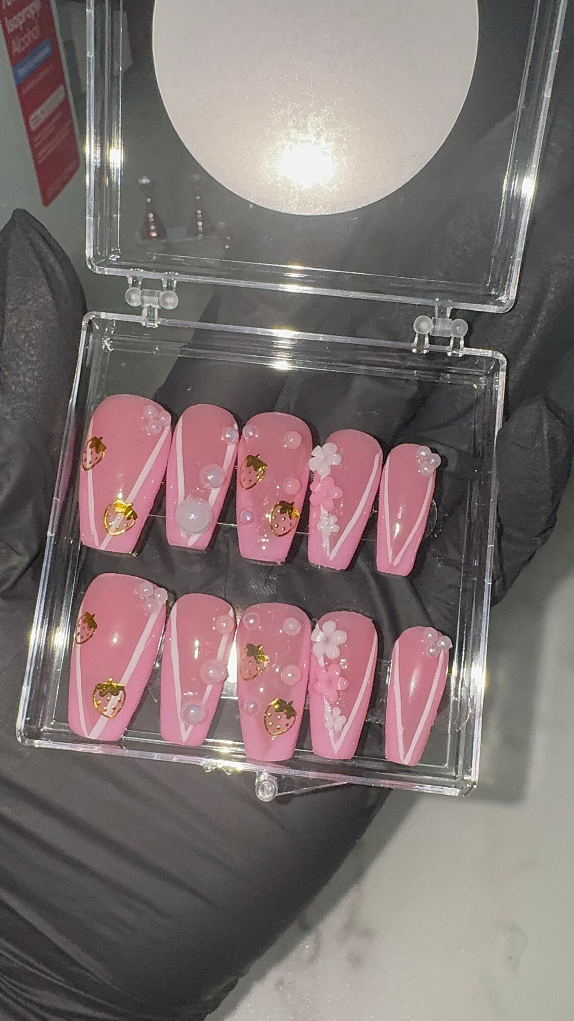 Pink false nails with white flower designs and gold accents in a clear plastic case, on a black surface.